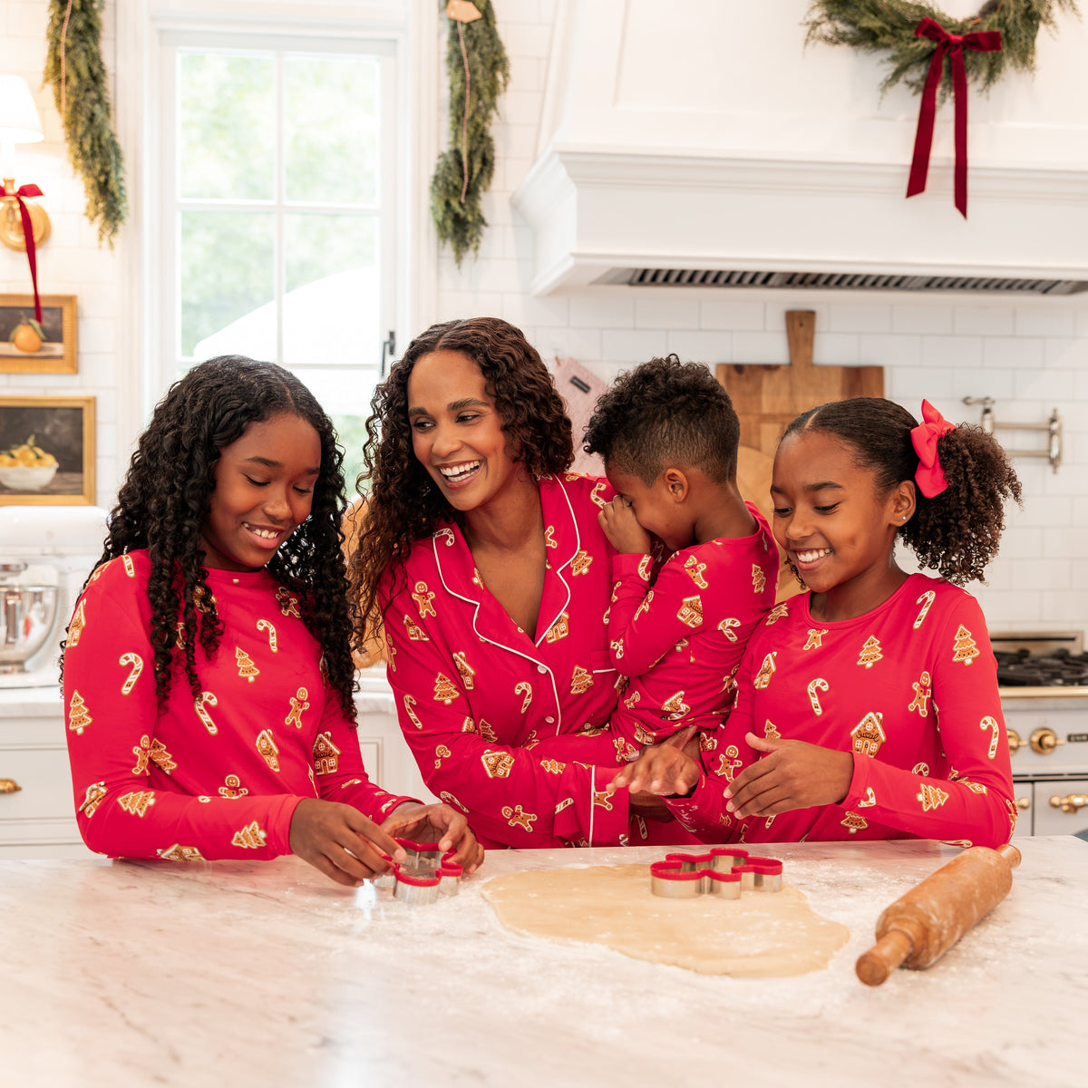 A family of four wearing matching red Christmas-themed pajamas is baking together in a bright white kitchen. The pajamas feature a festive gold pattern, and the children are using a rolling pin and cookie cutters on dough on the marble countertop.