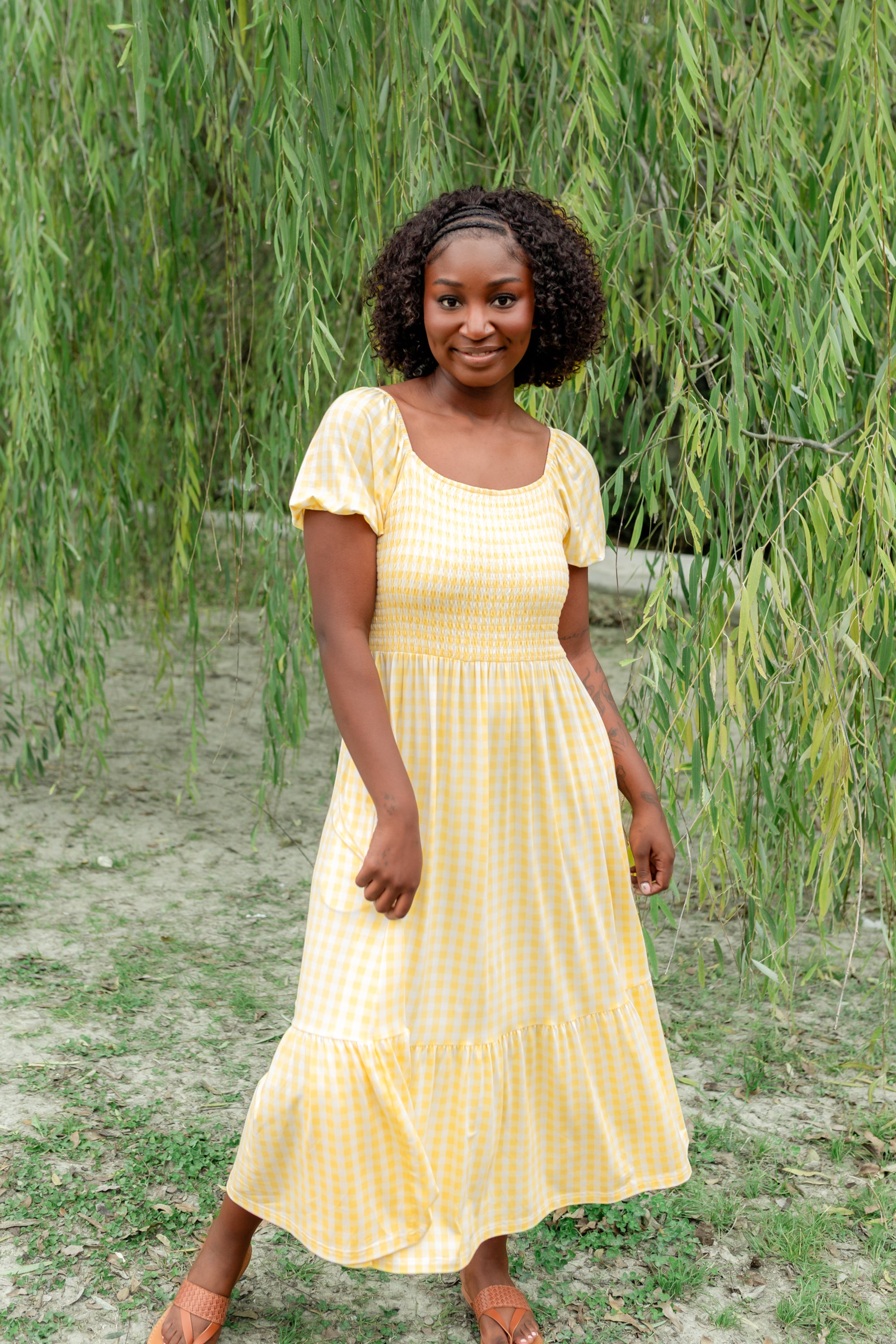 Smiling model standing in front of a tree wearing the Women’s Smocked Dress in Gingham Chamomile