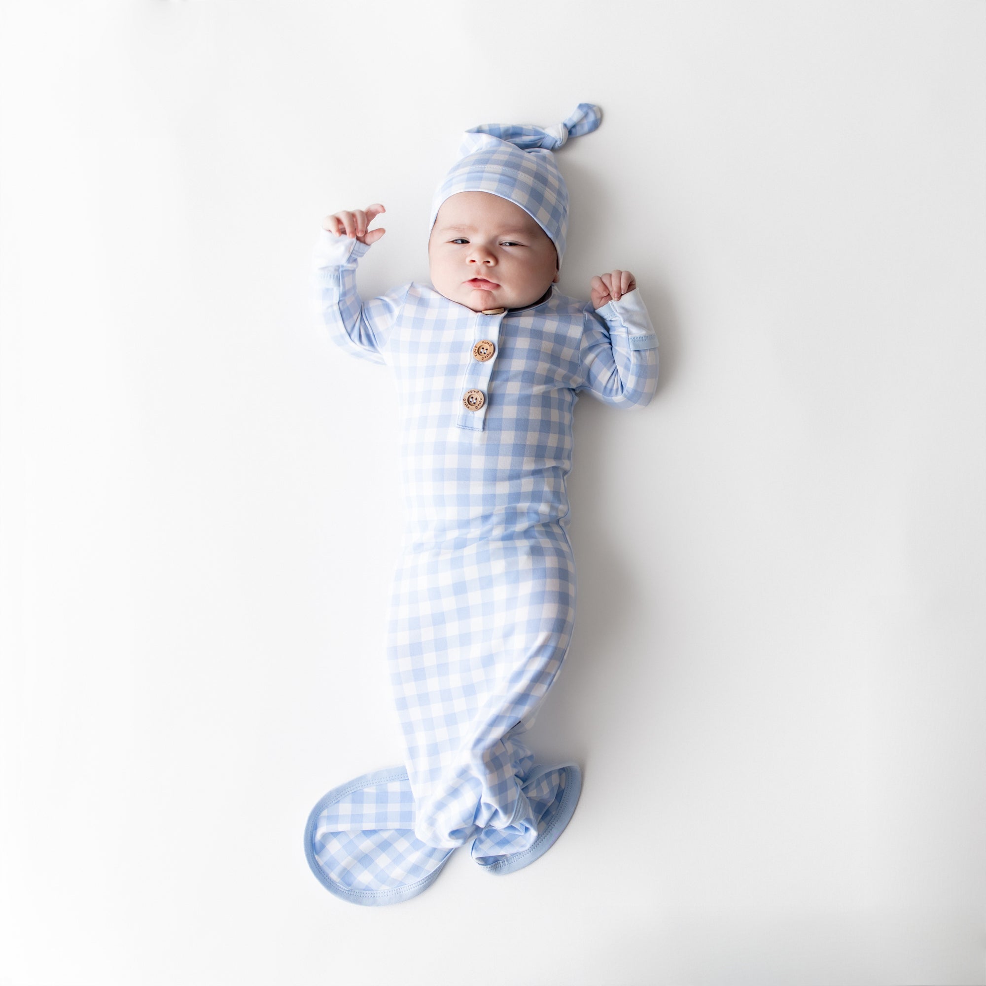 Newborn wearing the Knotted Gown with Hat Set in Gingham Breeze laying on a light cream surface