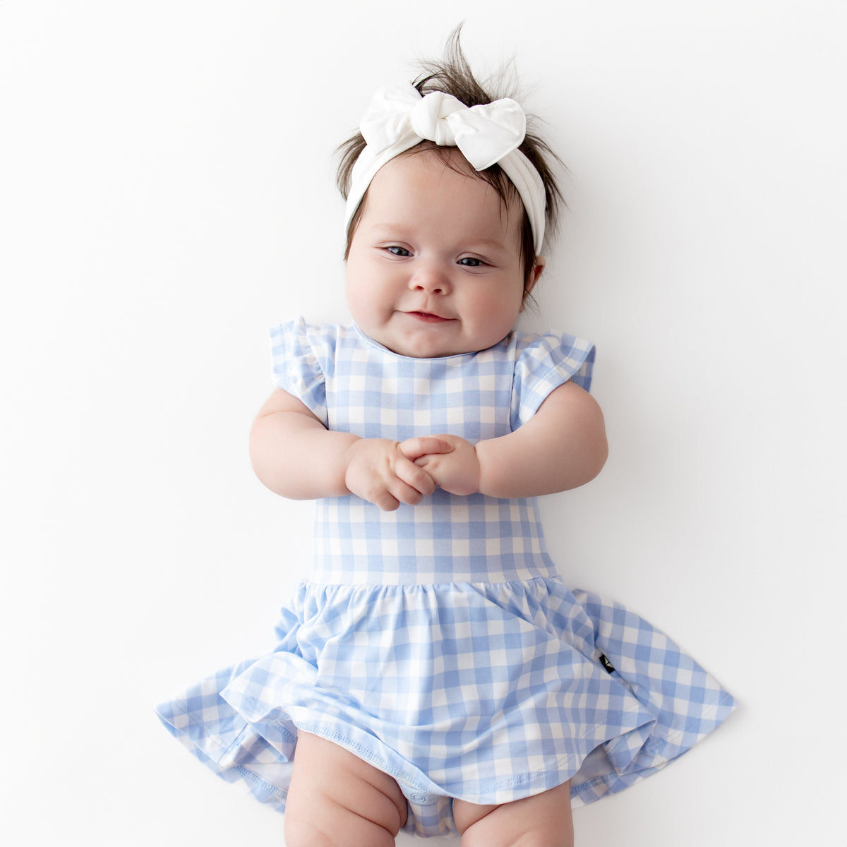 Infant laying on a light cream surface wearing the Twirl Bodysuit Dress in Gingham Breeze with a white knotted bow headband
