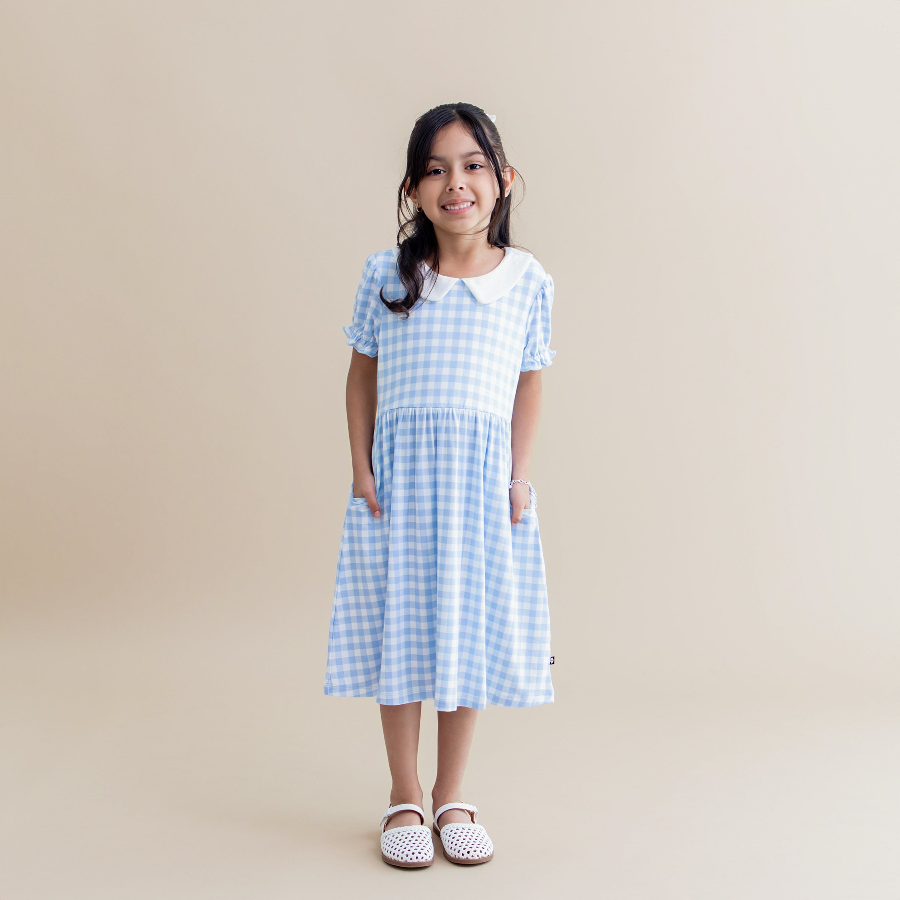 Young girl standing in front of a light cream surface wit her hands in the pocket of the Peter Pan Collar Dress in Gingham Breeze
