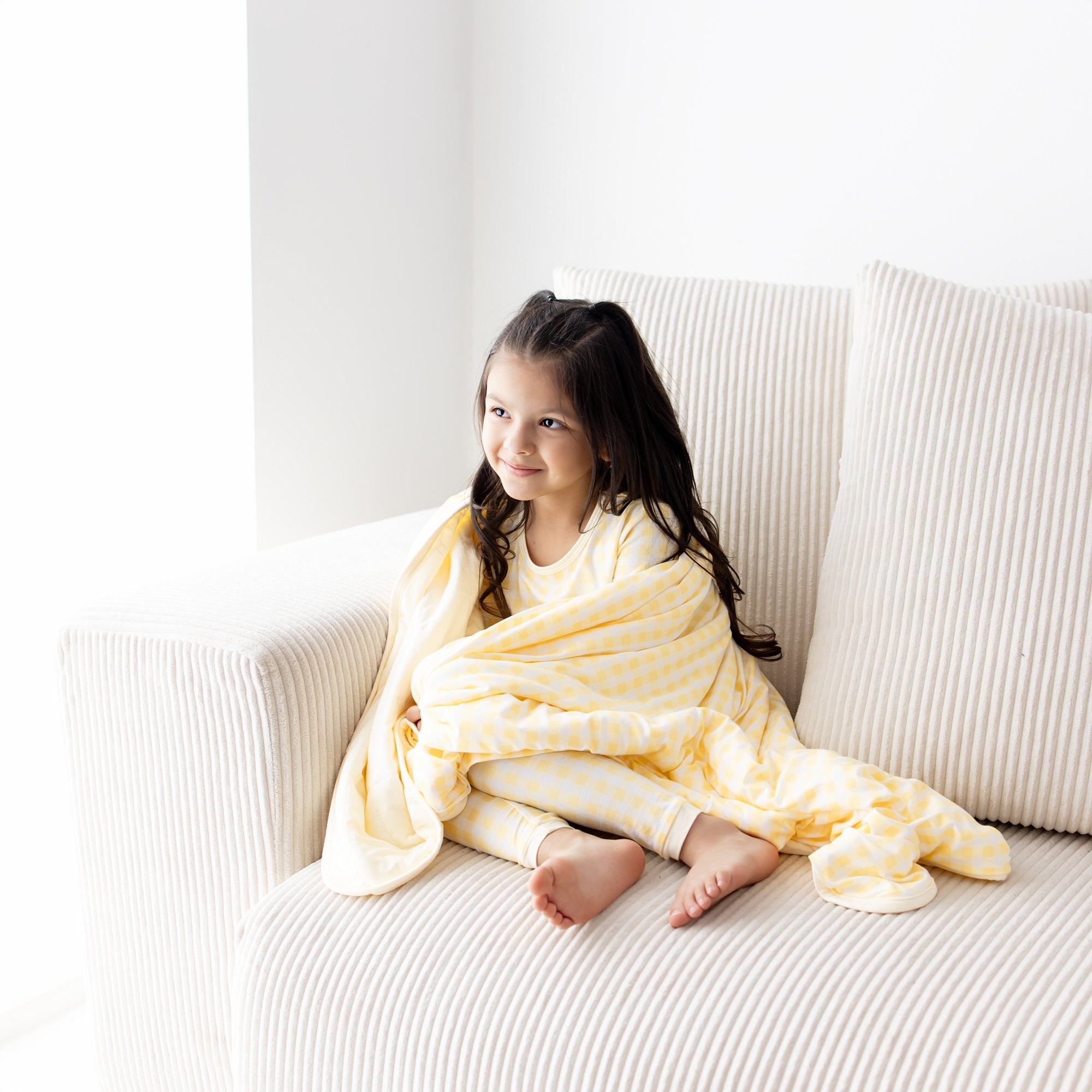 Young girl sitting on a couch with the Toddler Blanket in Gingham Chamomile 1.0 wrapped around her