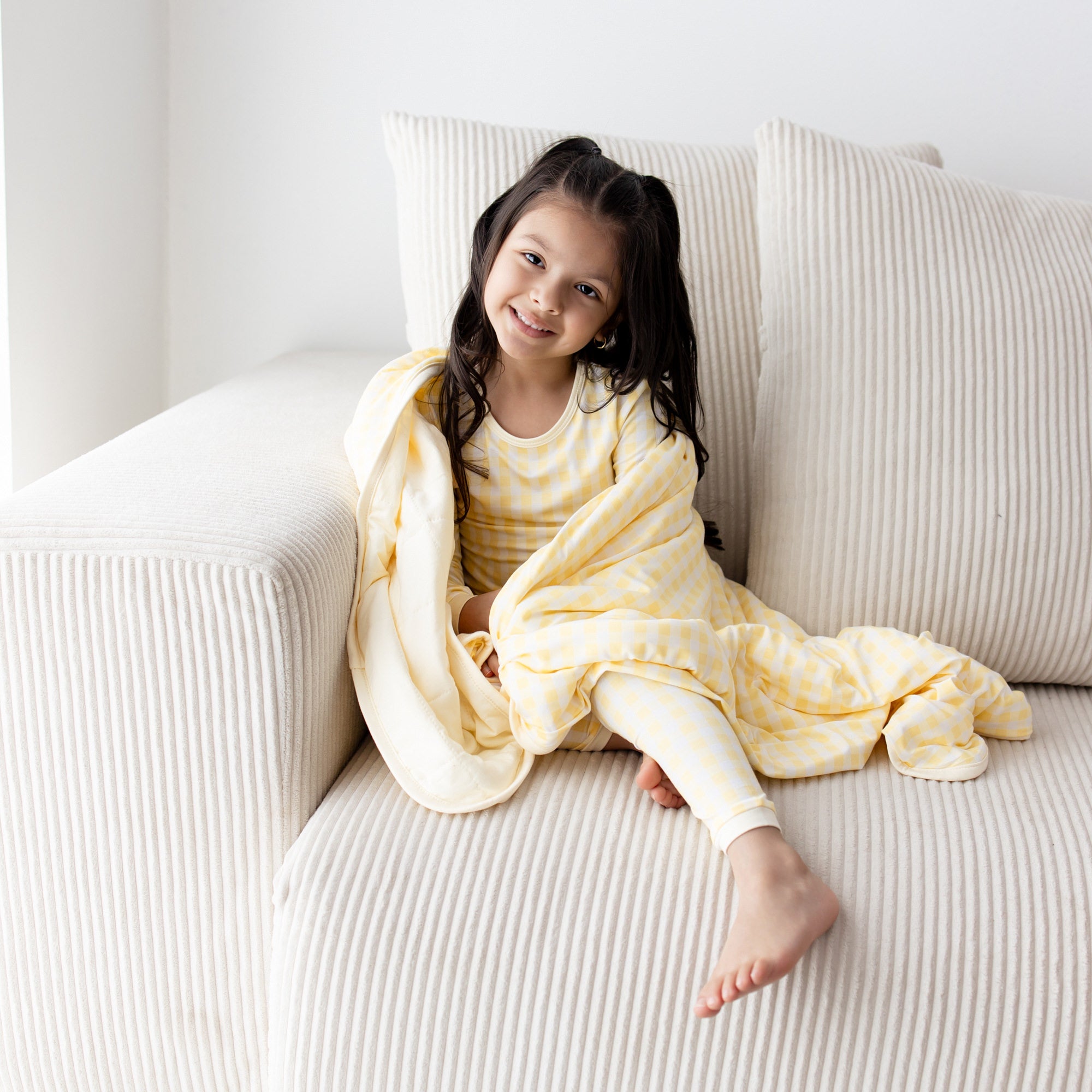 Young smiling girl sitting on a couch with the Toddler Blanket in Gingham Chamomile 1.0 around her shoulders wearing matching long sleeve pajamas