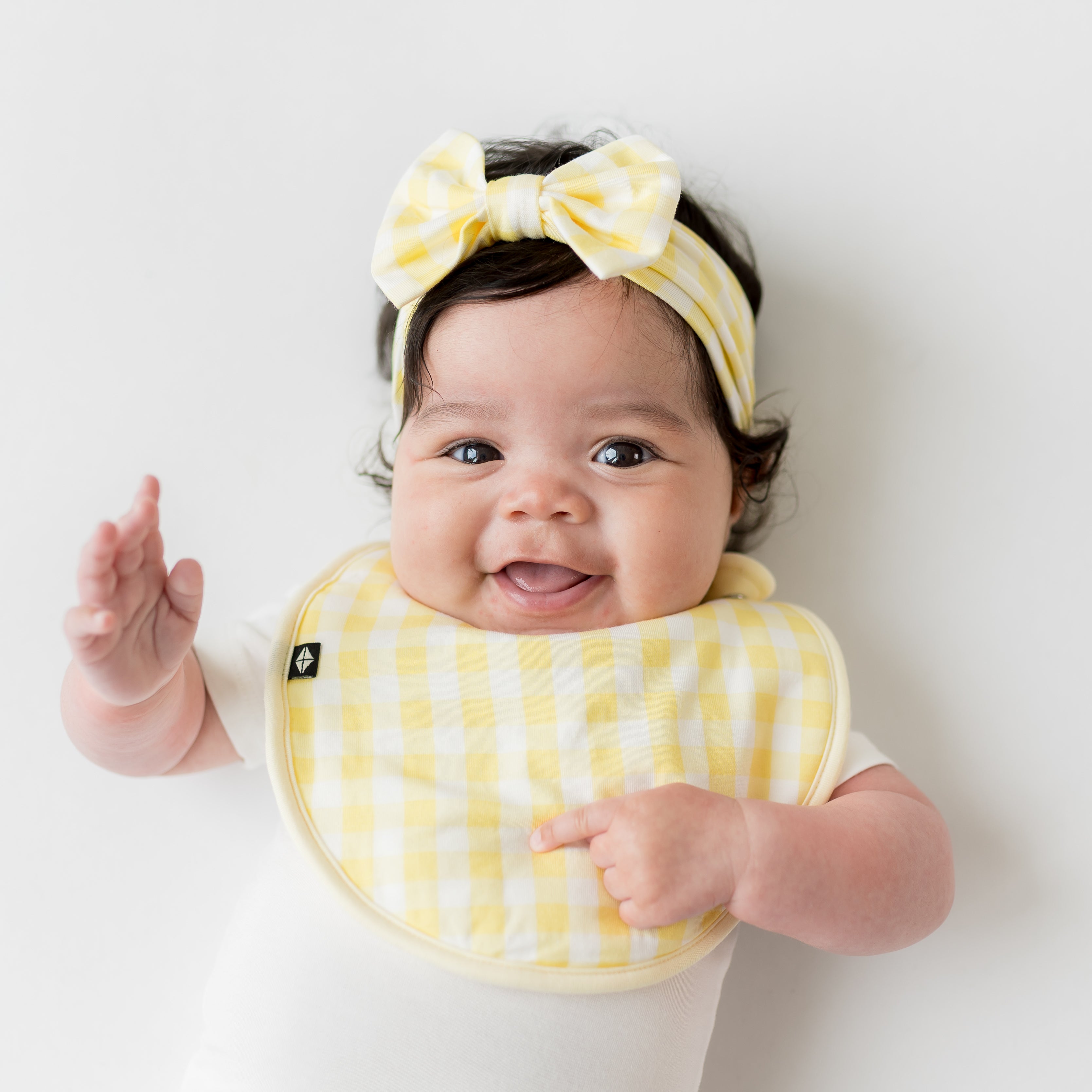 Smiling infant wearing the Cushy Bib in Gingham Chamomile and matching bow