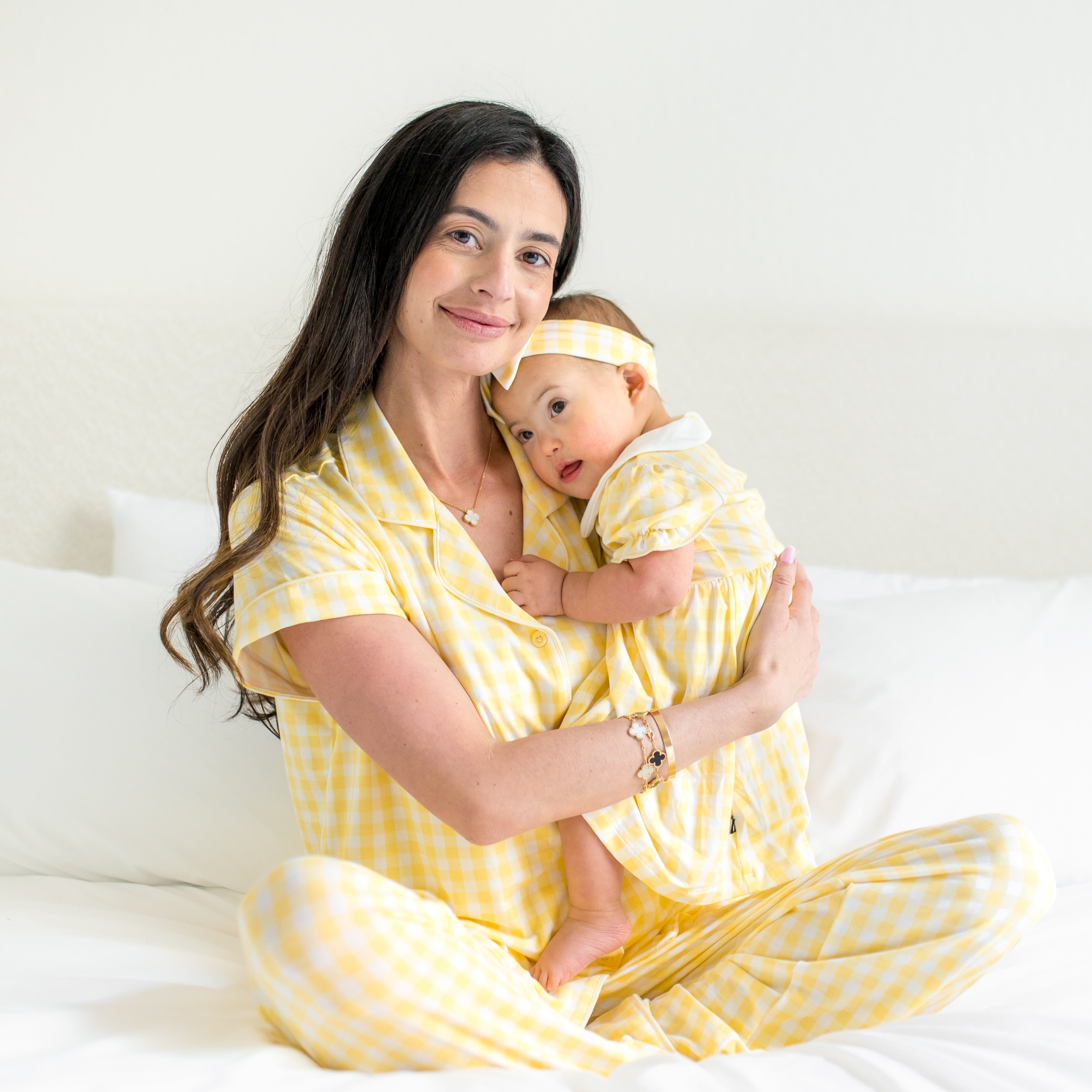 Young toddler wearing the Peter Pan Collar Bodysuit Dress in Gingham Chamomile being held by her mother who is sitting on a bed matching in women's pajamas