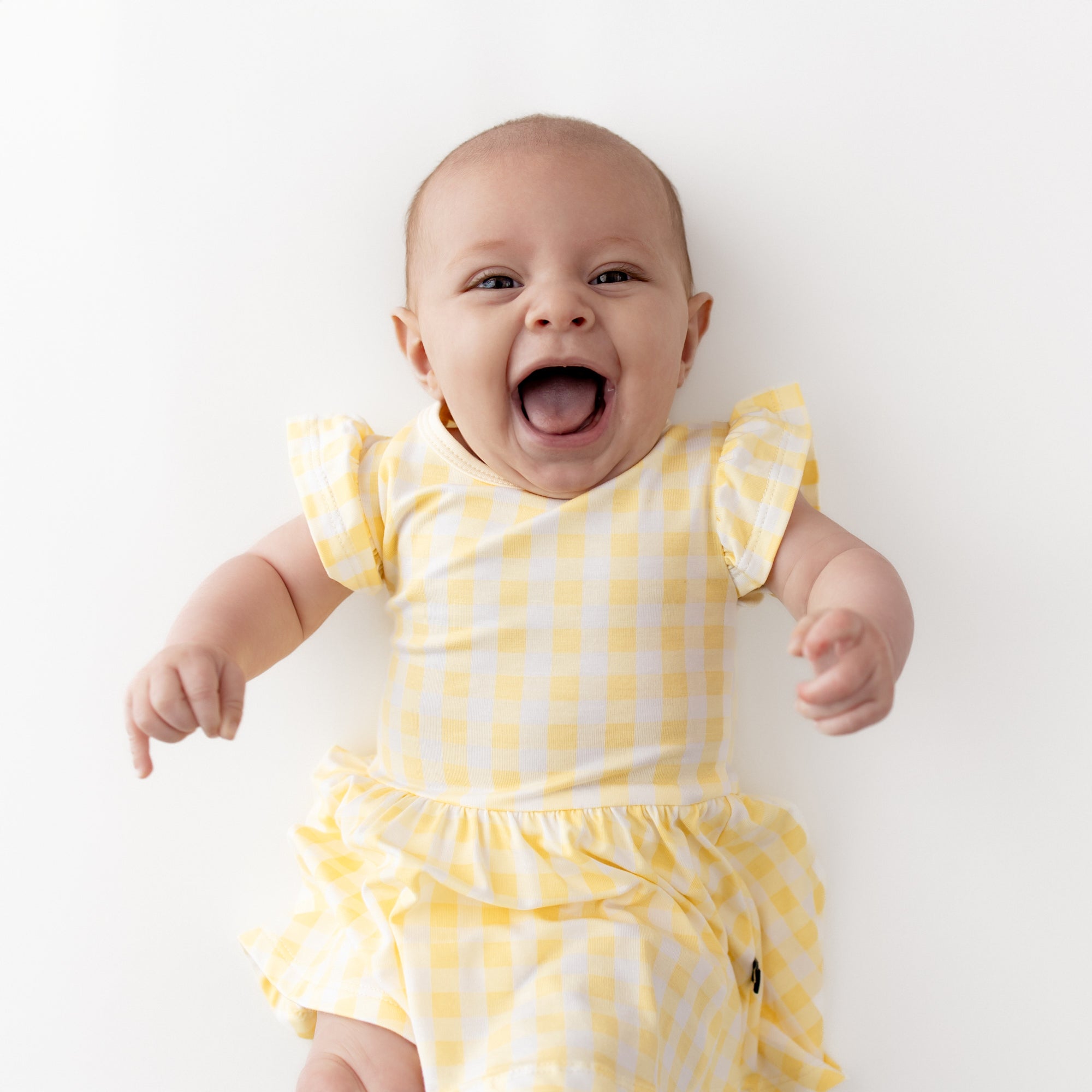 Smiling infant wearing the Twirl Bodysuit Dress in Gingham Chamomile laying on a light cream surface