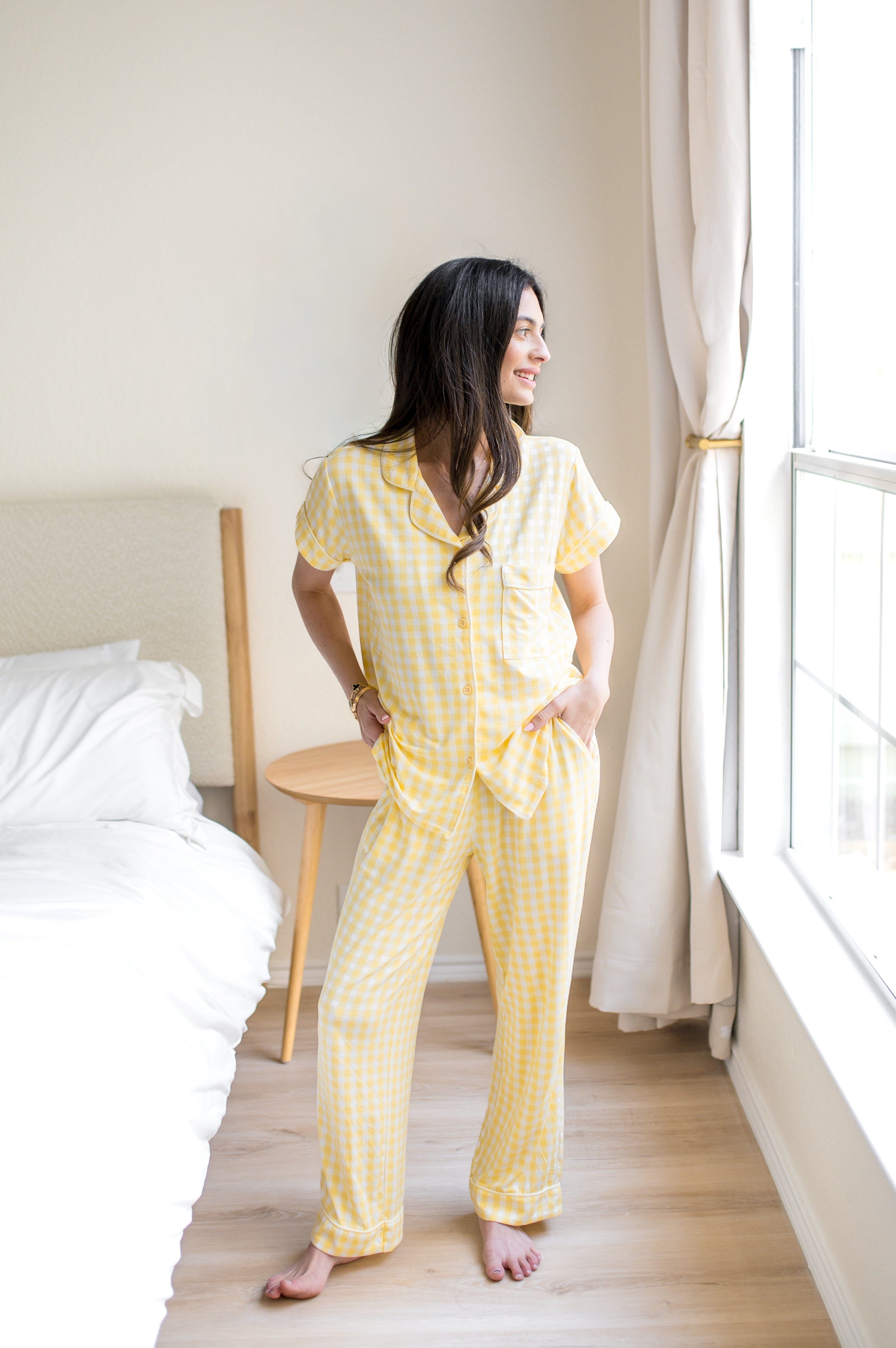 Model standing beside a bed wearing the Women's Short Sleeve Cropped Pajama Set in Gingham Chamomile