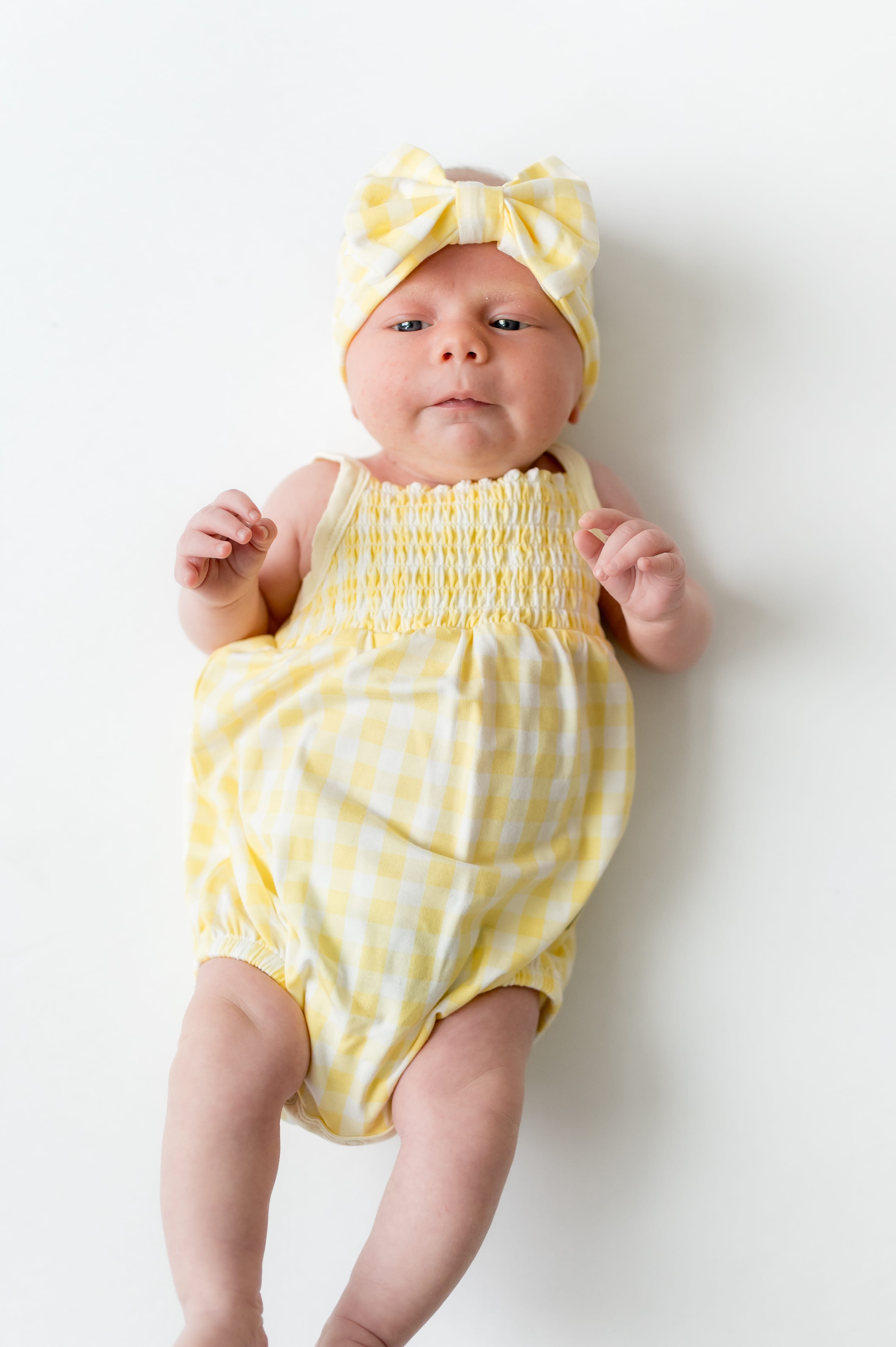 Newborn laying on a white surface wearing the Smocked Bubble Romper in Gingham Chamomile with matching bow