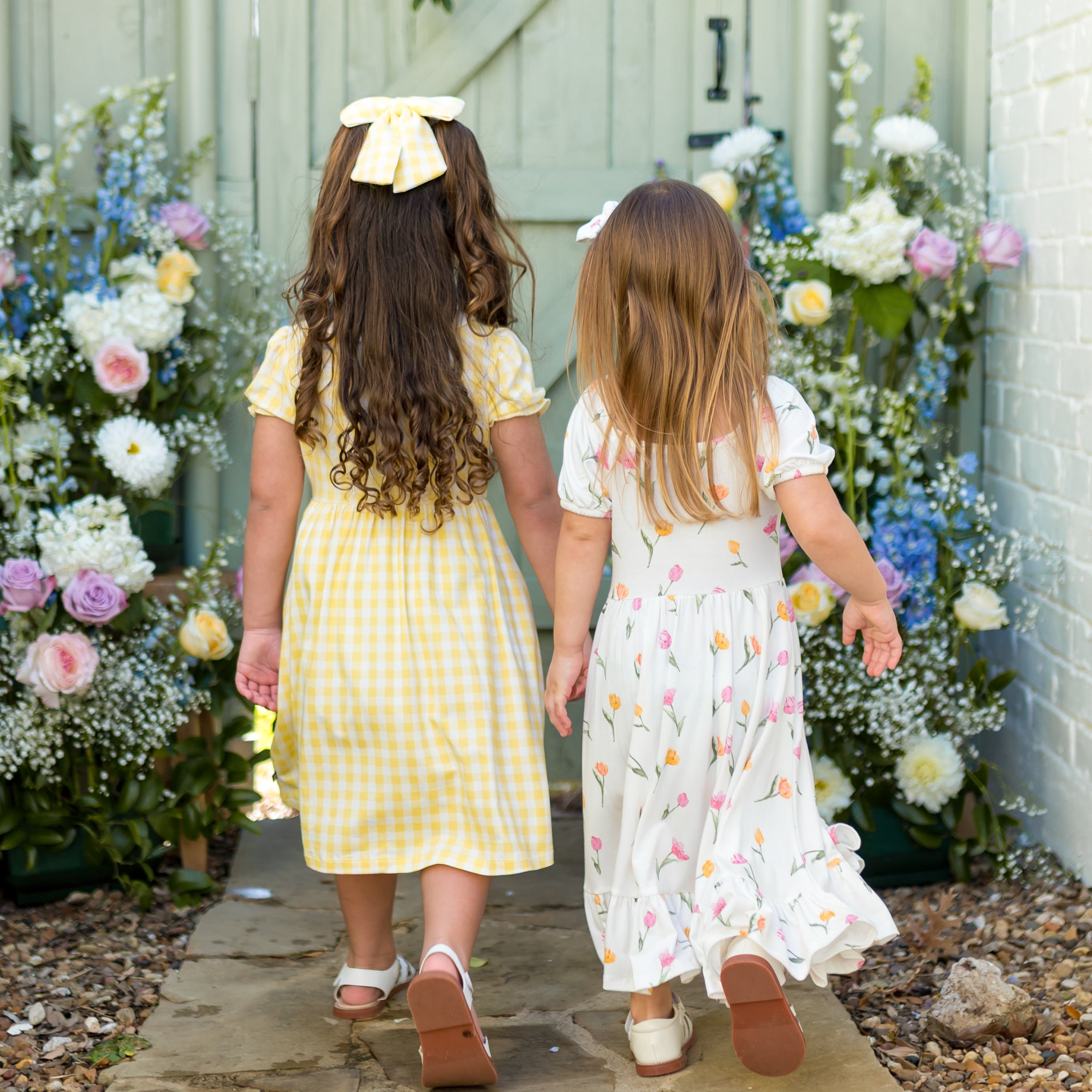 Two girls walking down a stone path away from the camera towards a green fence and flowers showing the back of the Peter Pan Collar Dress in Gingham Chamomile and Puff Sleeve Twirl Dress in Tulip
