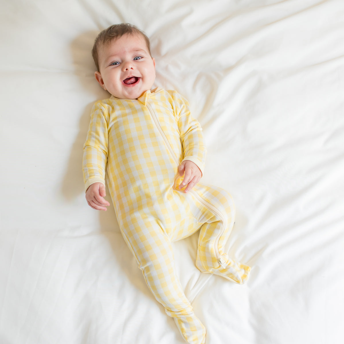 Young infant laying in a bed wearing the Zippered Footie in Gingham Chamomile