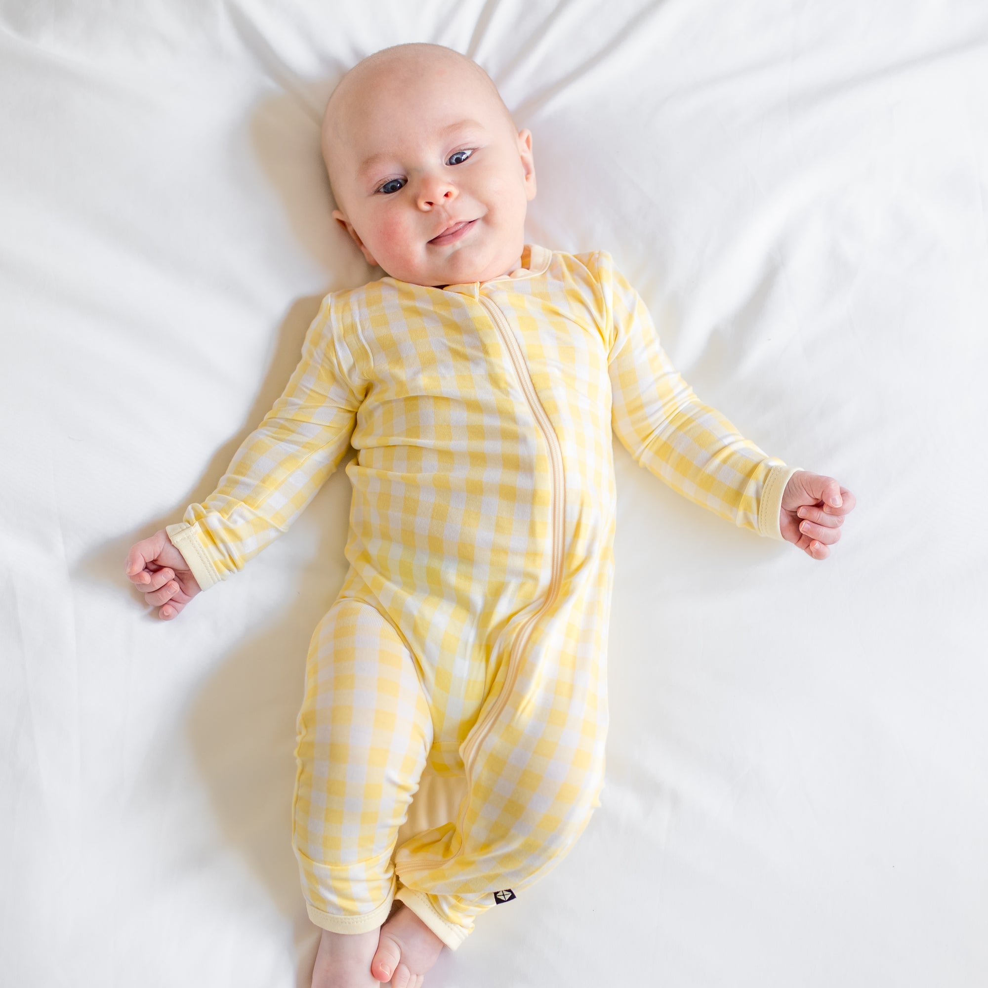 Young infant laying in bed wearing the Zippered Romper in Gingham Chamomile