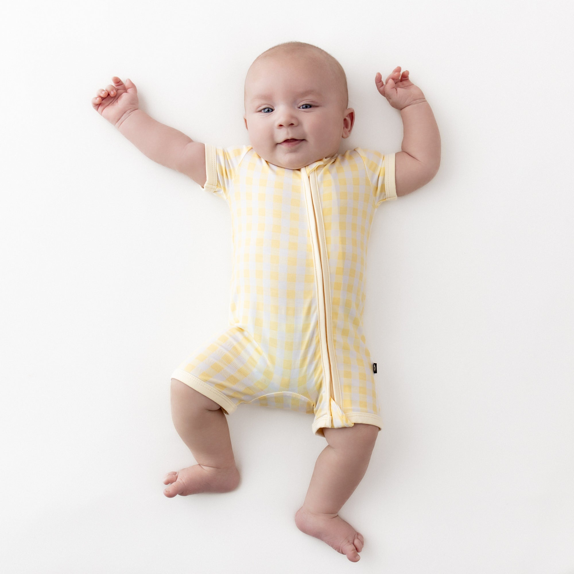 Infant wearing the Zipper Shortall Romper in Gingham Chamomile stretched out on a light neutral surface