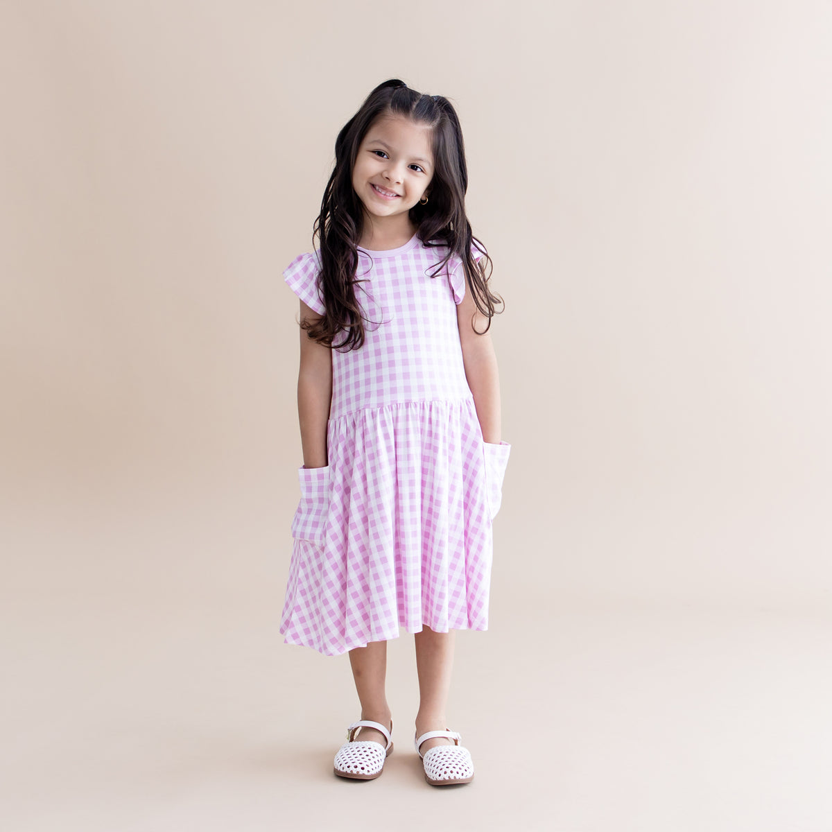 Young girl posing with her hands in the pocket of the Pocket Dress in Gingham Thistle with white sandals on