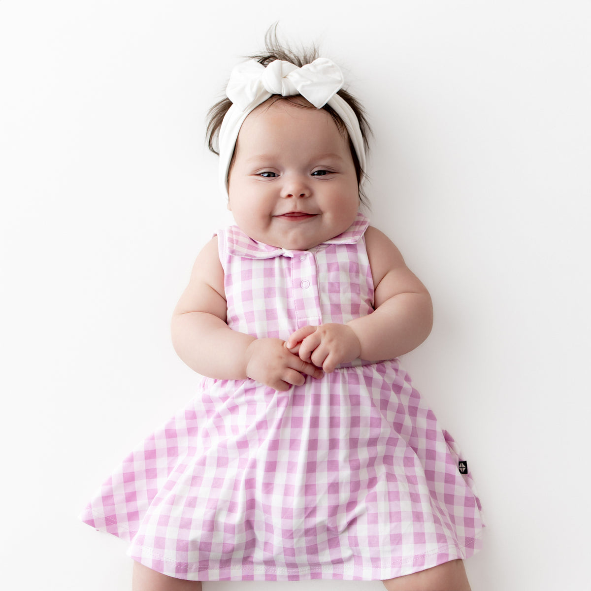 Young infant wearing the Polo Bodysuit Dress in Gingham Thistle with a white knotted bow headband