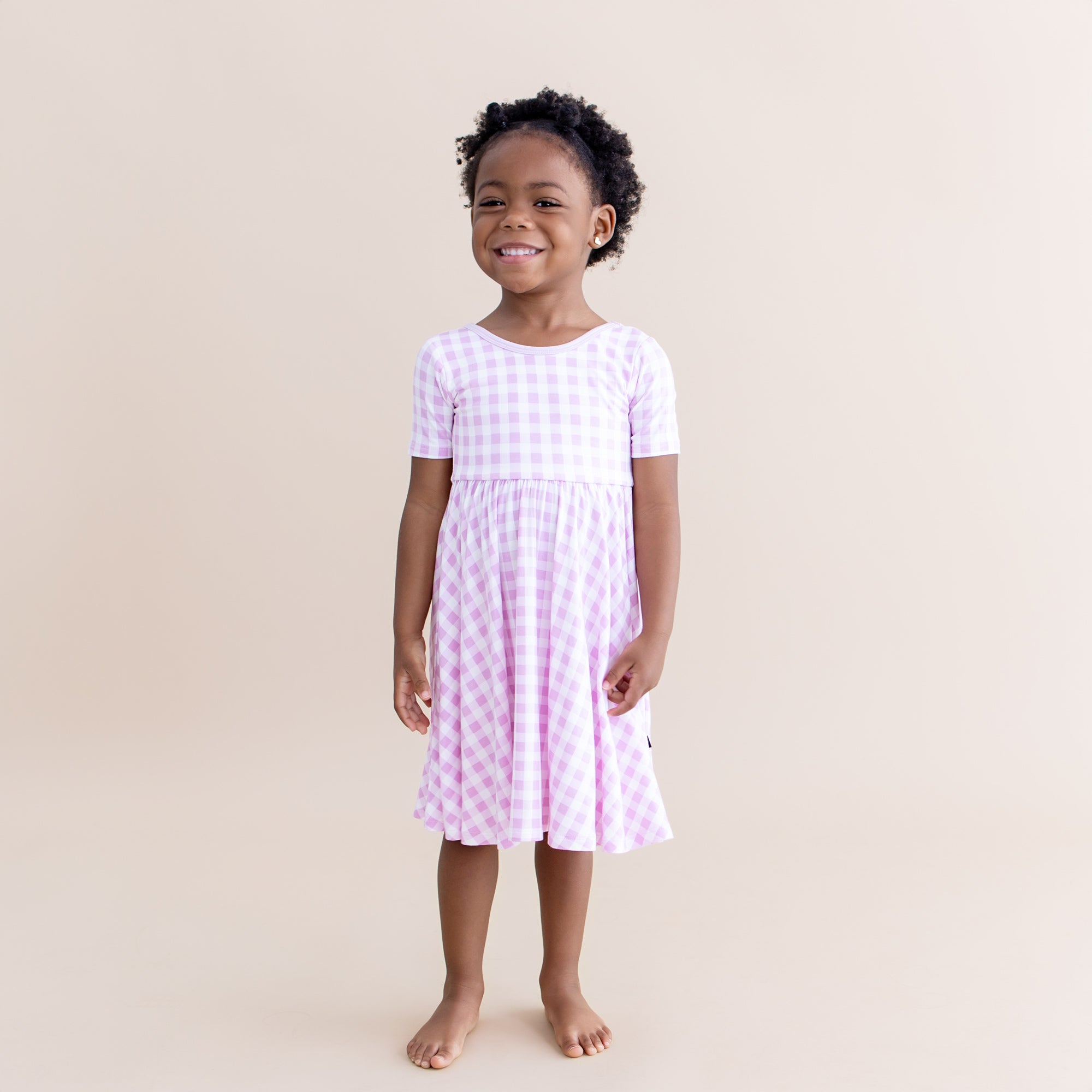 Young girl smiling wearing the Twirl Dress in Gingham Thistle standing in front of a light beige background