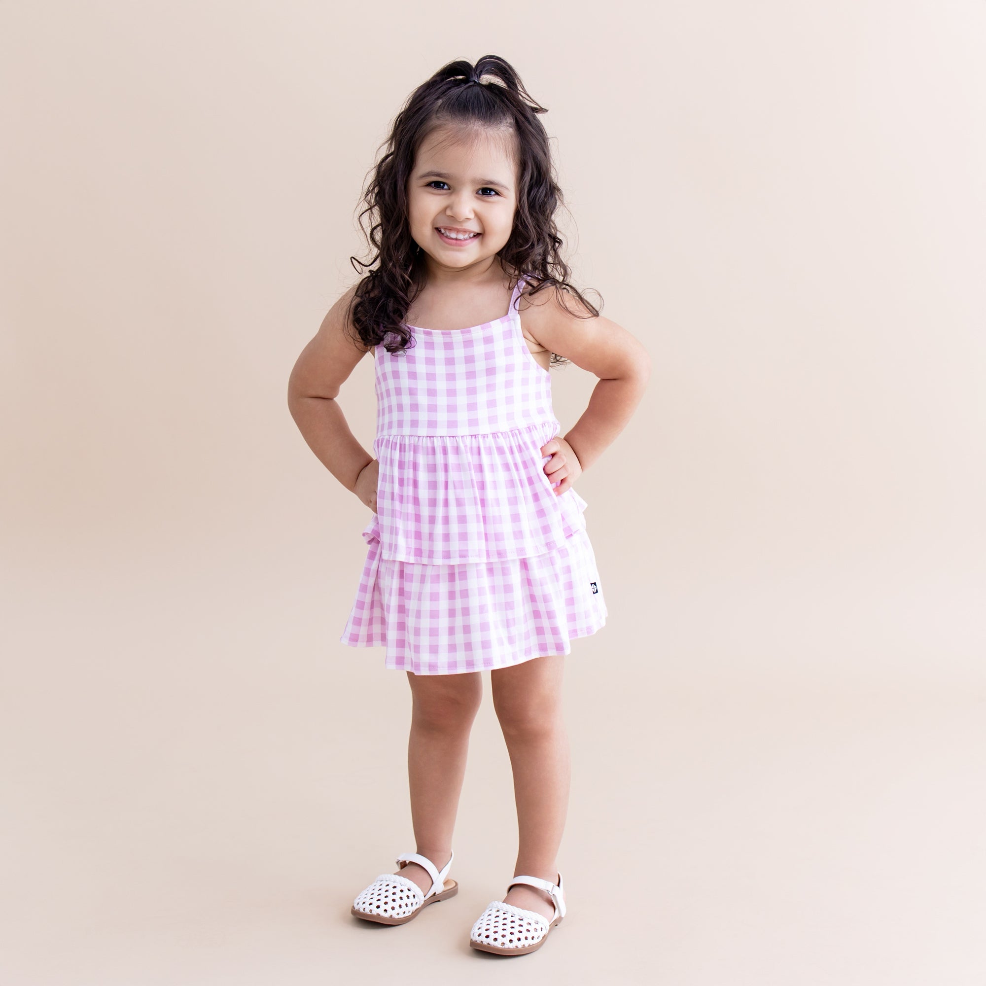 Toddler posing wearing the Spaghetti Strap Ruffle Romper in Gingham Thistle paired with white sandals