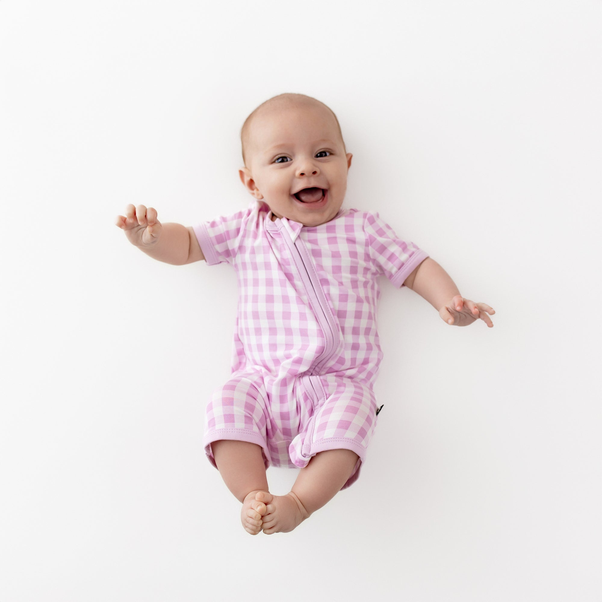 Smiling infant wearing the Zipper Shortall Romper in Gingham Thistle laying on a light cream surface