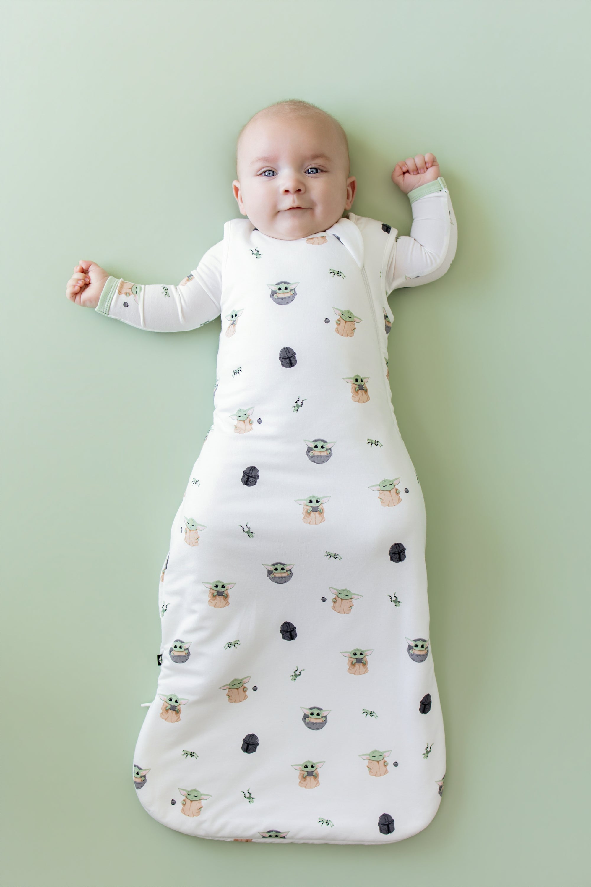 Infant laying on a light green surface wearing the Sleep Bag in Grogu™ 1.0 with a matching long sleeve romper on underneath