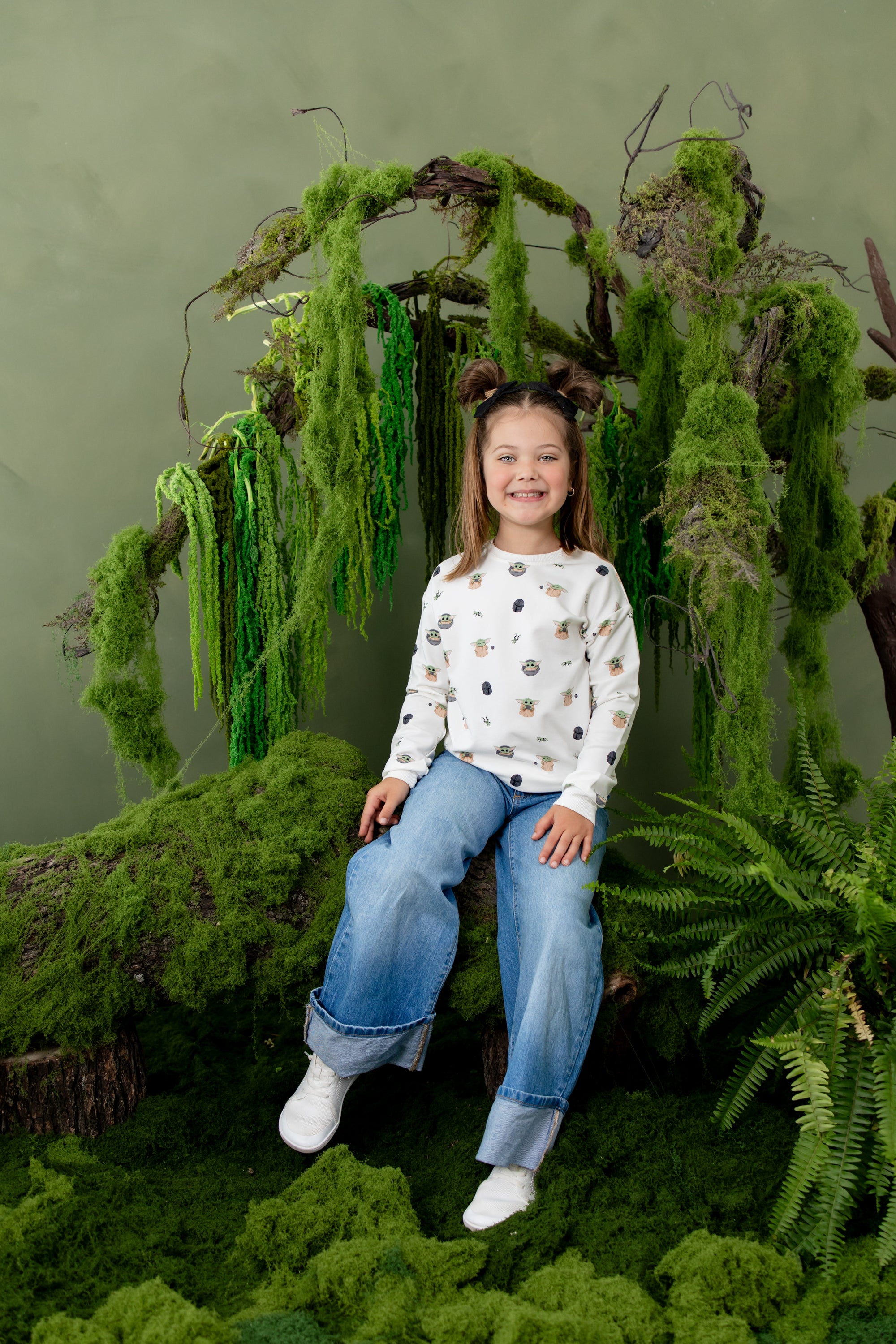 Young girl sitting on a tree branch with moss on it wearing the Sweatshirt in Grogu™ and medium wash jeans
