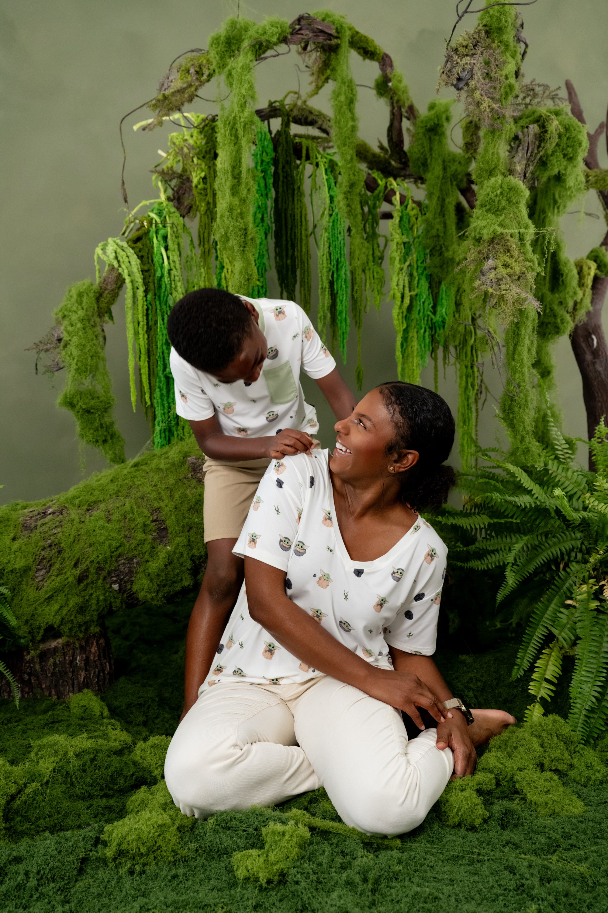 Female sitting on the ground wearing the Women's Relaxed Fit V-Neck in Grogu™ with her son standing behind him wearing matching crew neck tee both in front of greenery