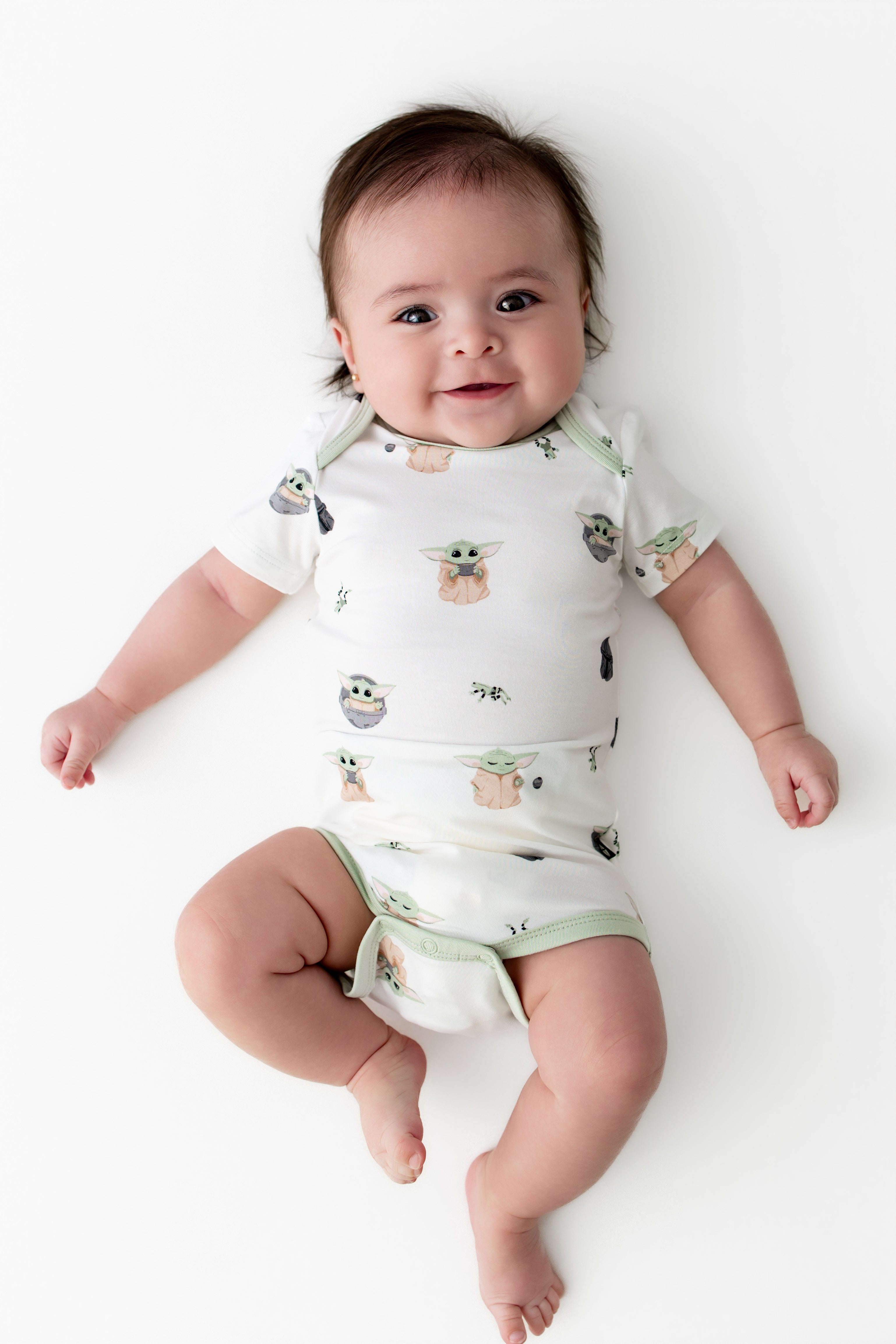Smiling infant laying on a white surface wearing the Bodysuit in Grogu™