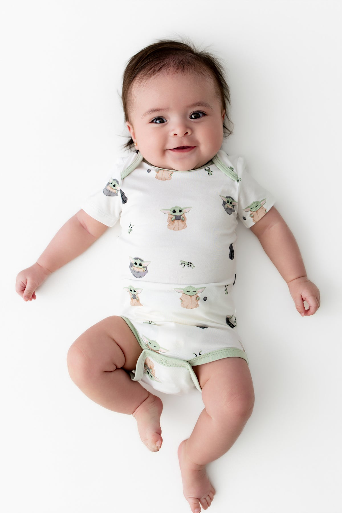 Smiling infant laying on a white surface wearing the Bodysuit in Grogu™