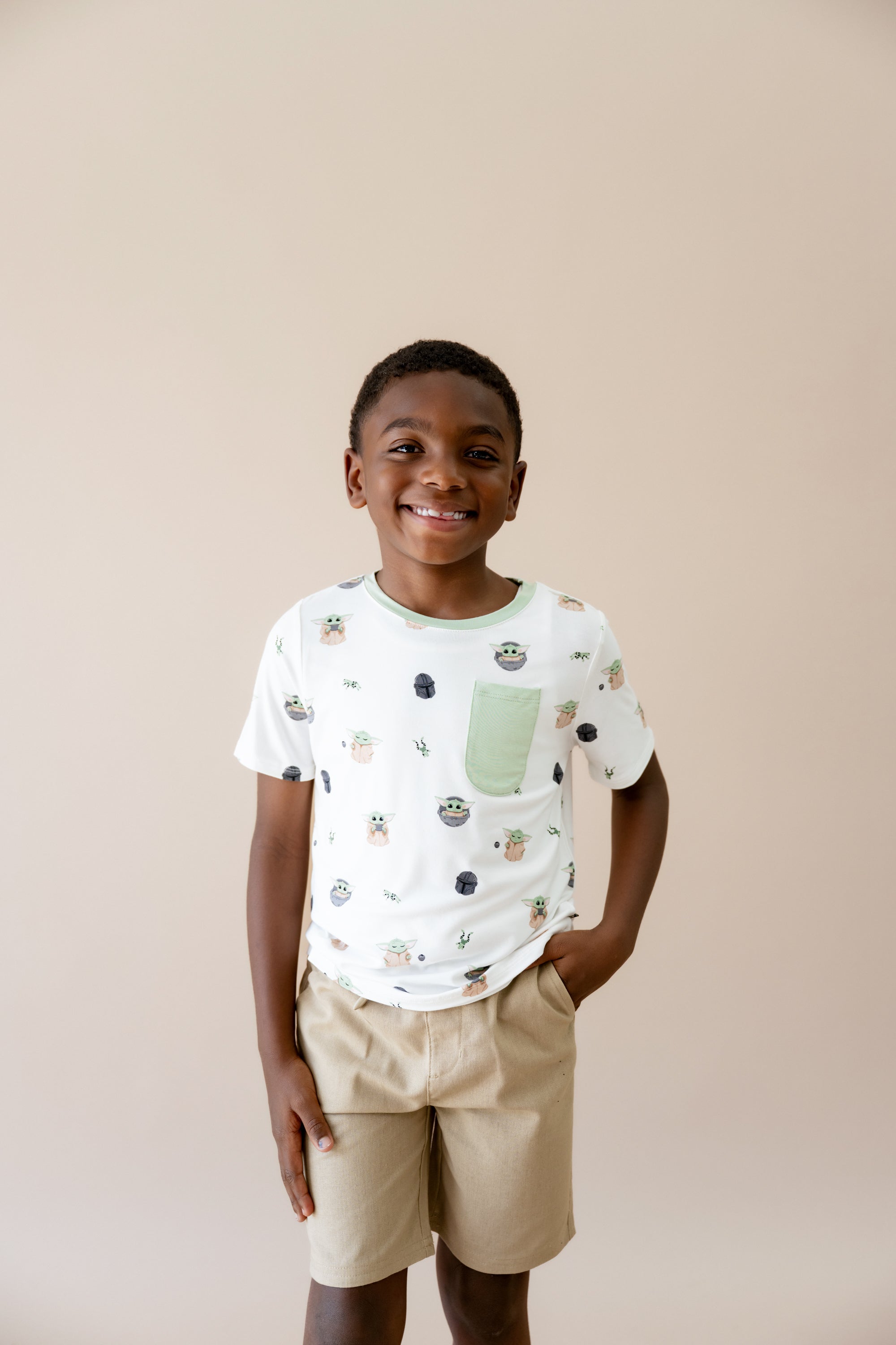 Smiling boy standing in front of a taupe background wearing the Toddler Crew Neck Tee in Grogu™ with tan shorts