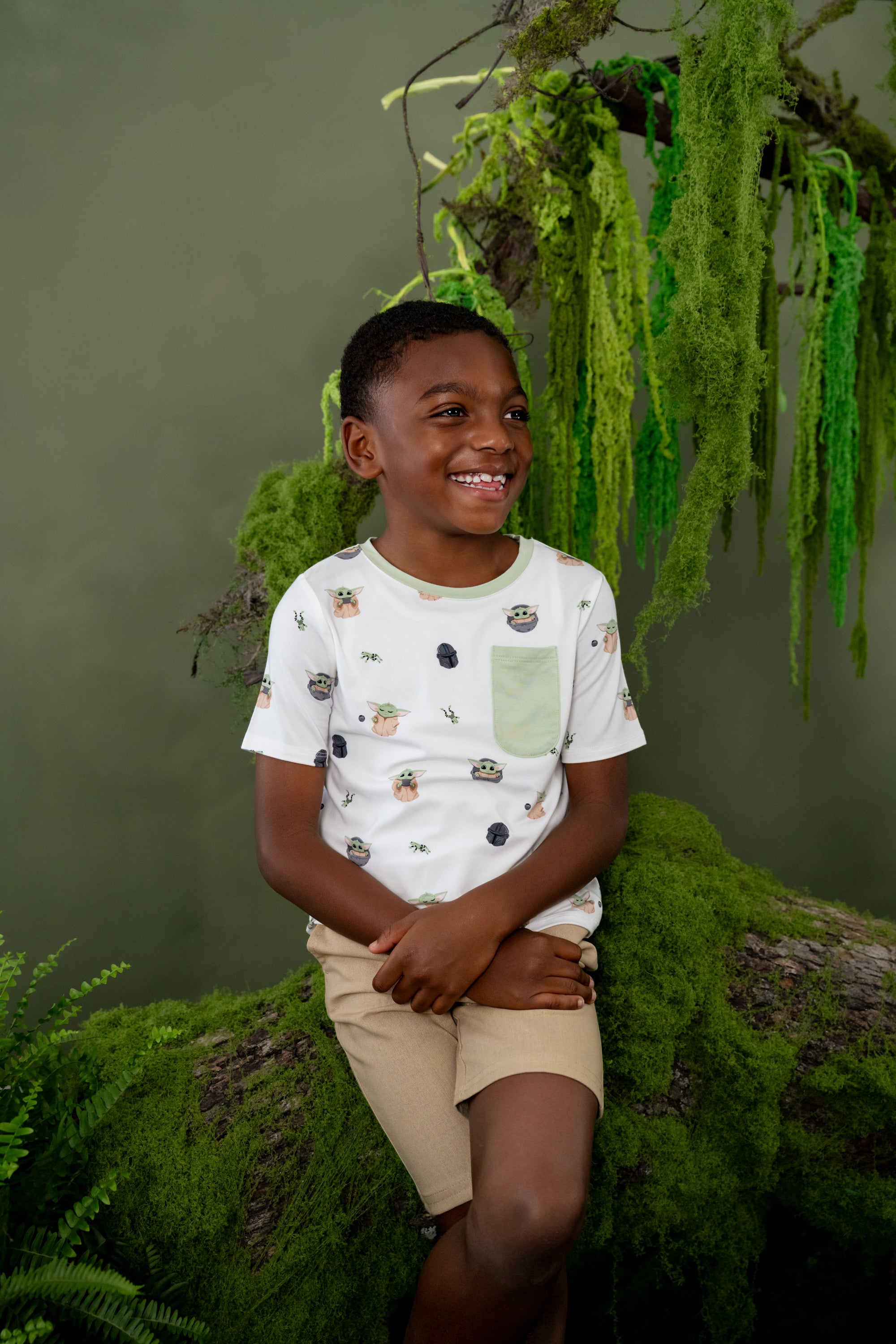 Smiling boy sitting on a tree branch with Moss on it wearing the Toddler Crew Neck Tee in Grogu™