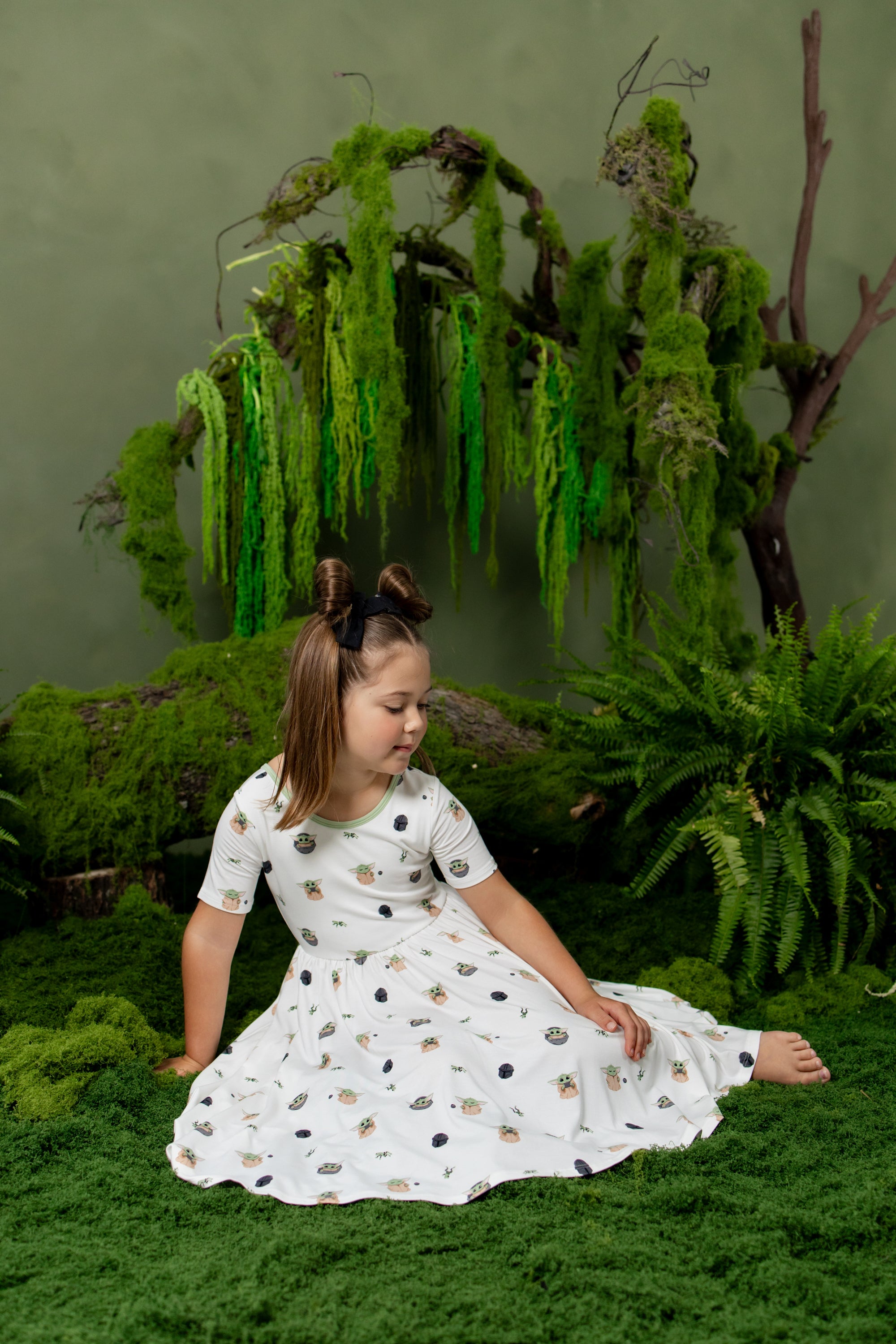 Young girl sitting on moss in front of trees and greenery wearing the Twirl Dress in Grogu™