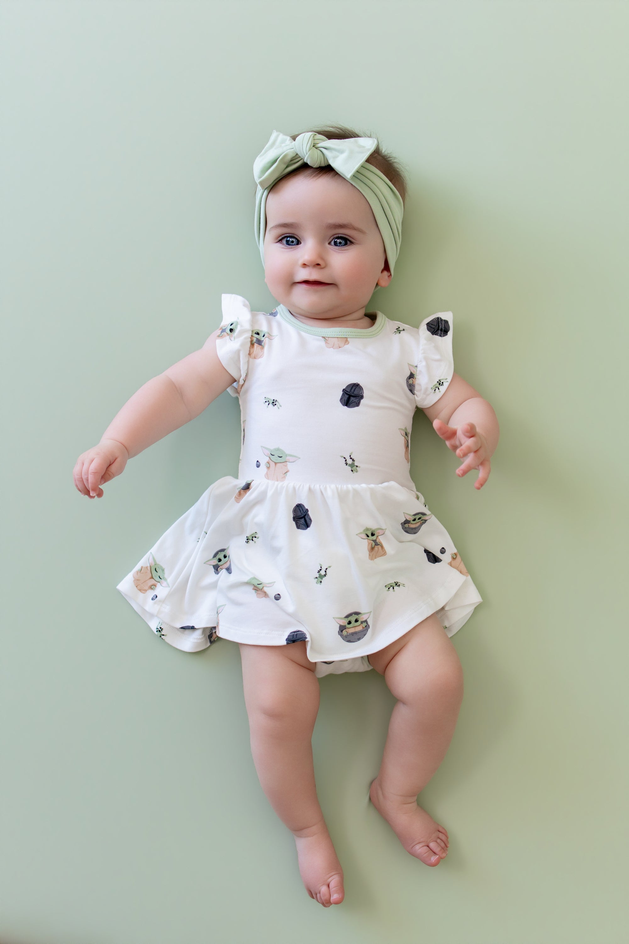Infant girl laying on a light green surface wearing the Twirl Bodysuit Dress in Grogu™ with a light green bow