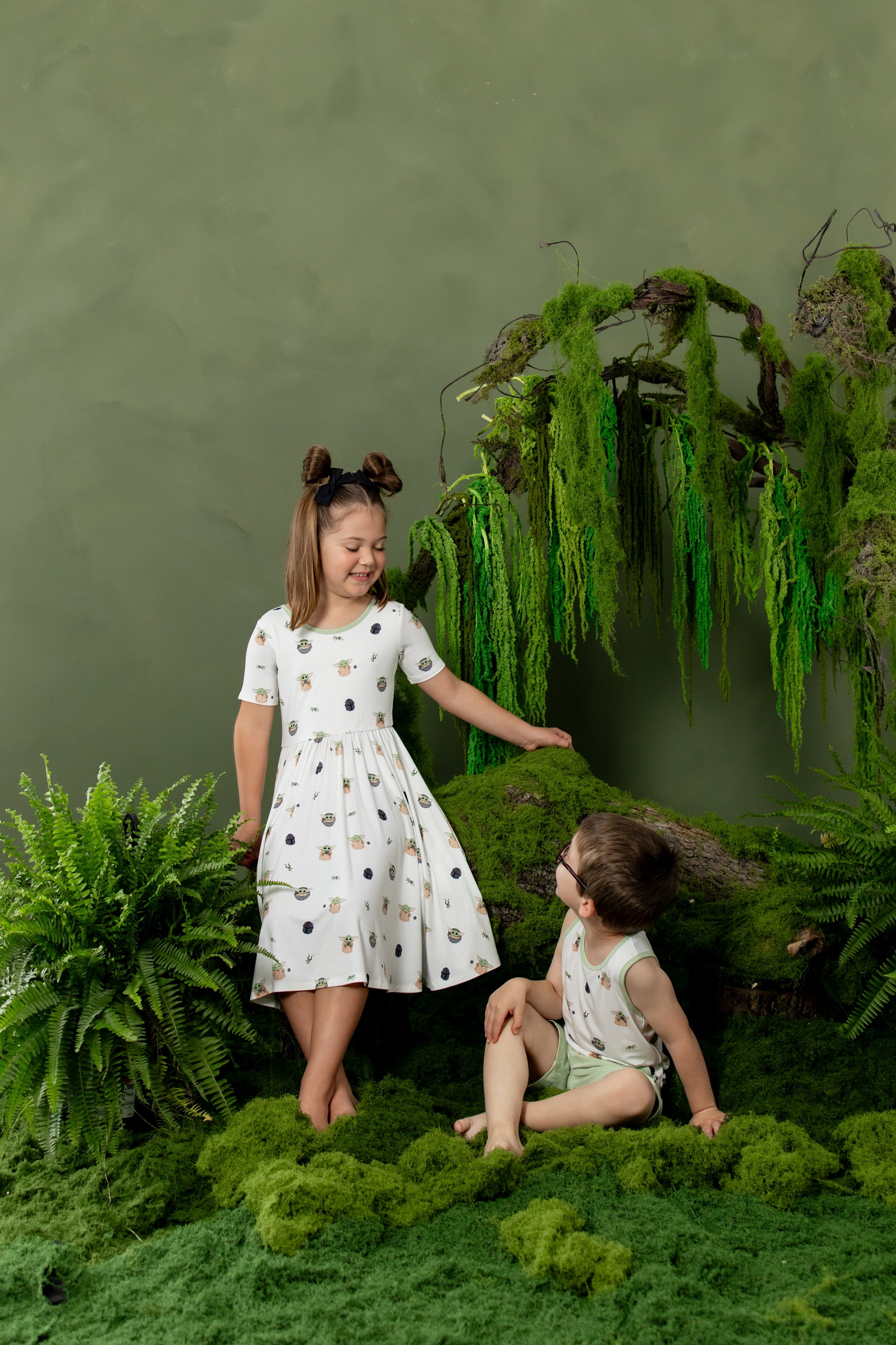 Young girl wearing the Twirl Dress in Grogu™ standing beside a young boy wearing the matching tank top with a greenery backdrop