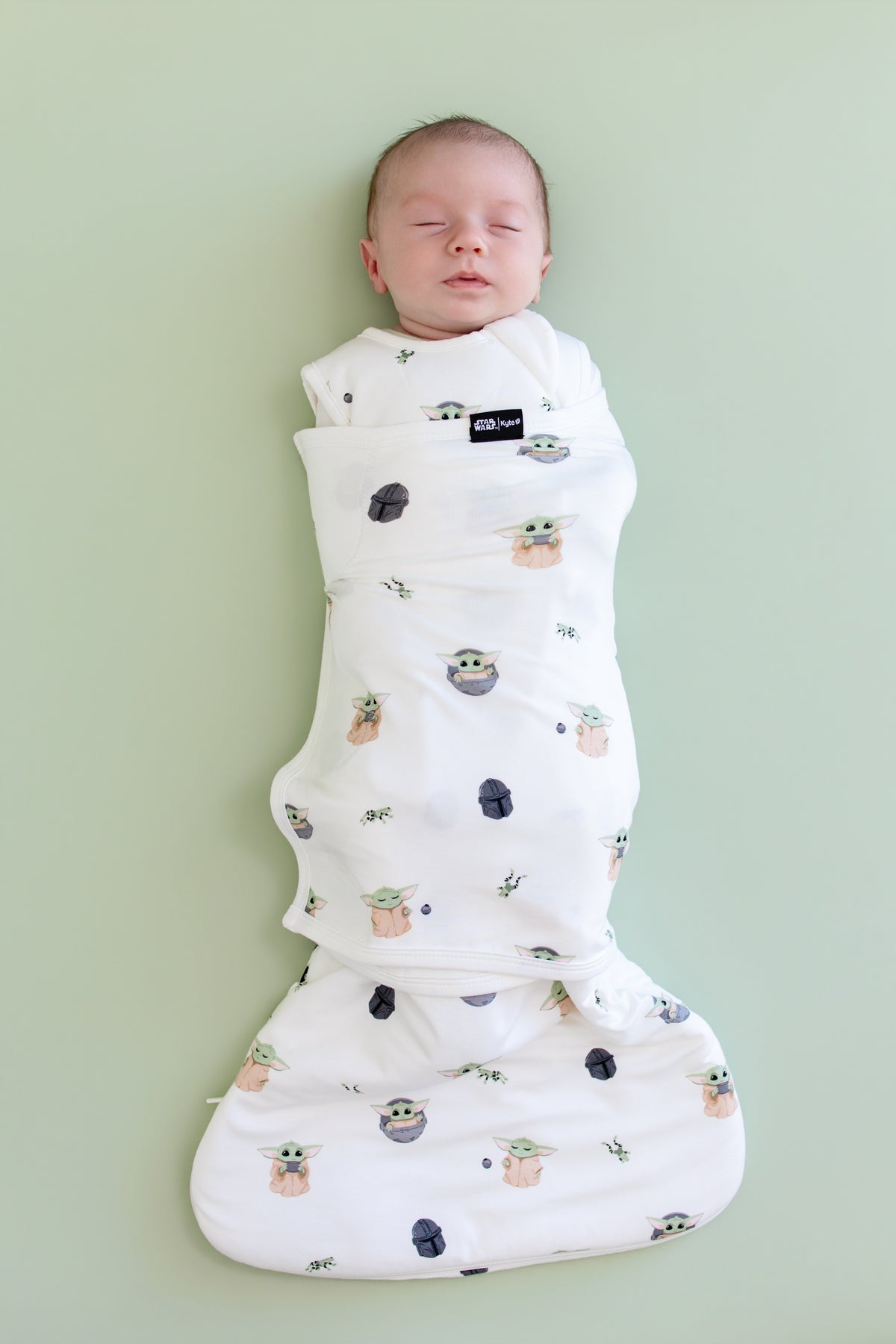 Sleeping newborn swaddled in the Sleep Bag Swaddler in Grogu™ 1.0 on a light green background