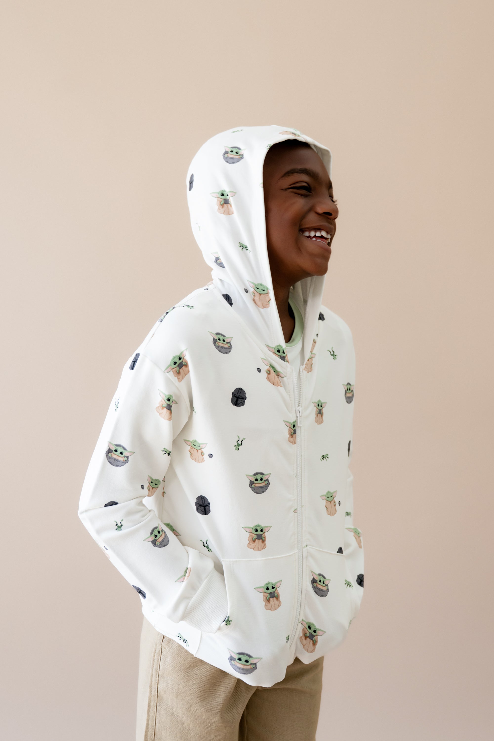 Young boy smiling looking off to the side wearing the Zip-Up Hoodie in Grogu™ with the hood up and his hands in the pockets