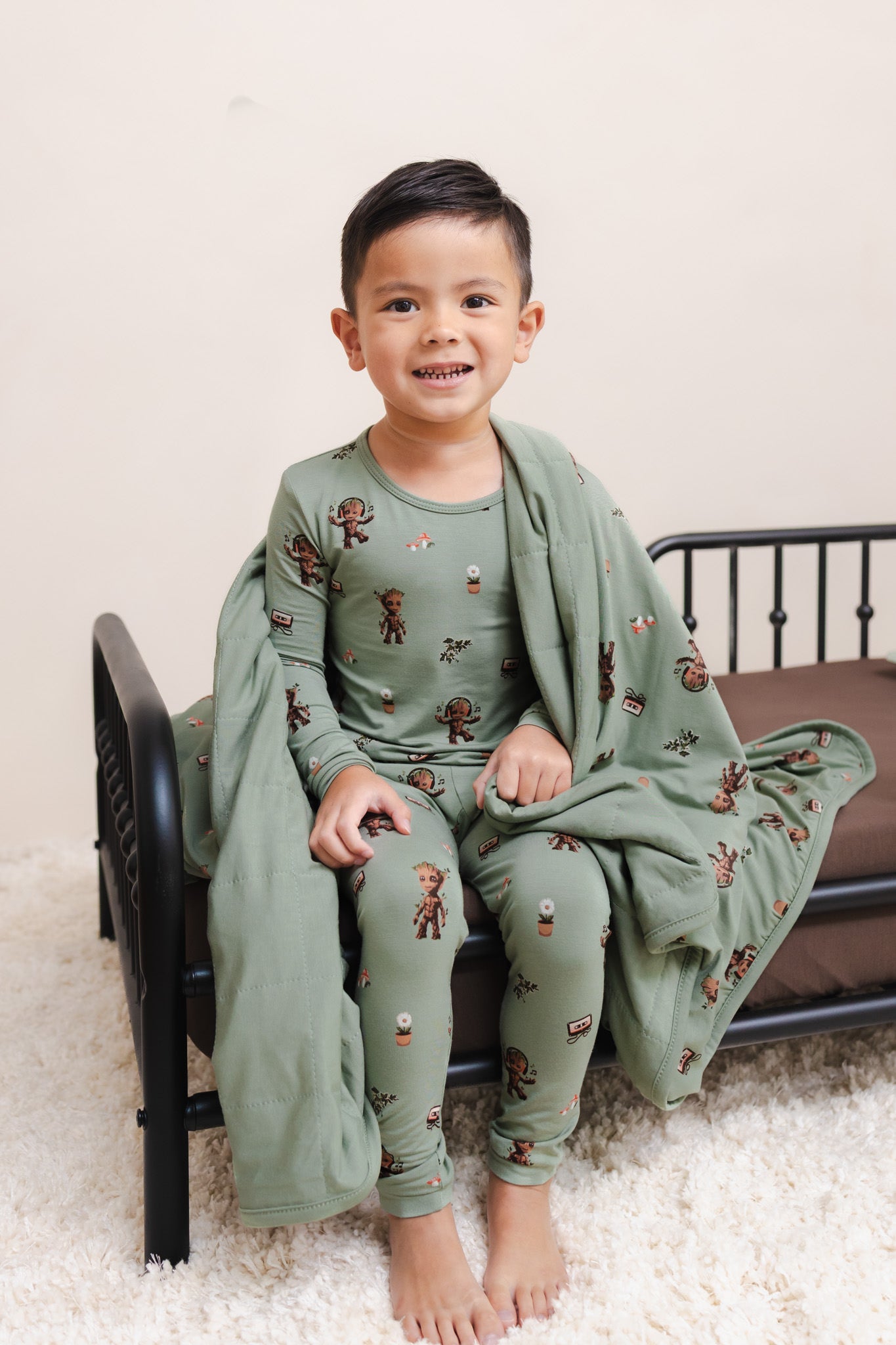 Boy wrapped in Green Toddler Blanket in Groot 1.0 print sitting on a bed wearing a similar print long sleeve pajama set