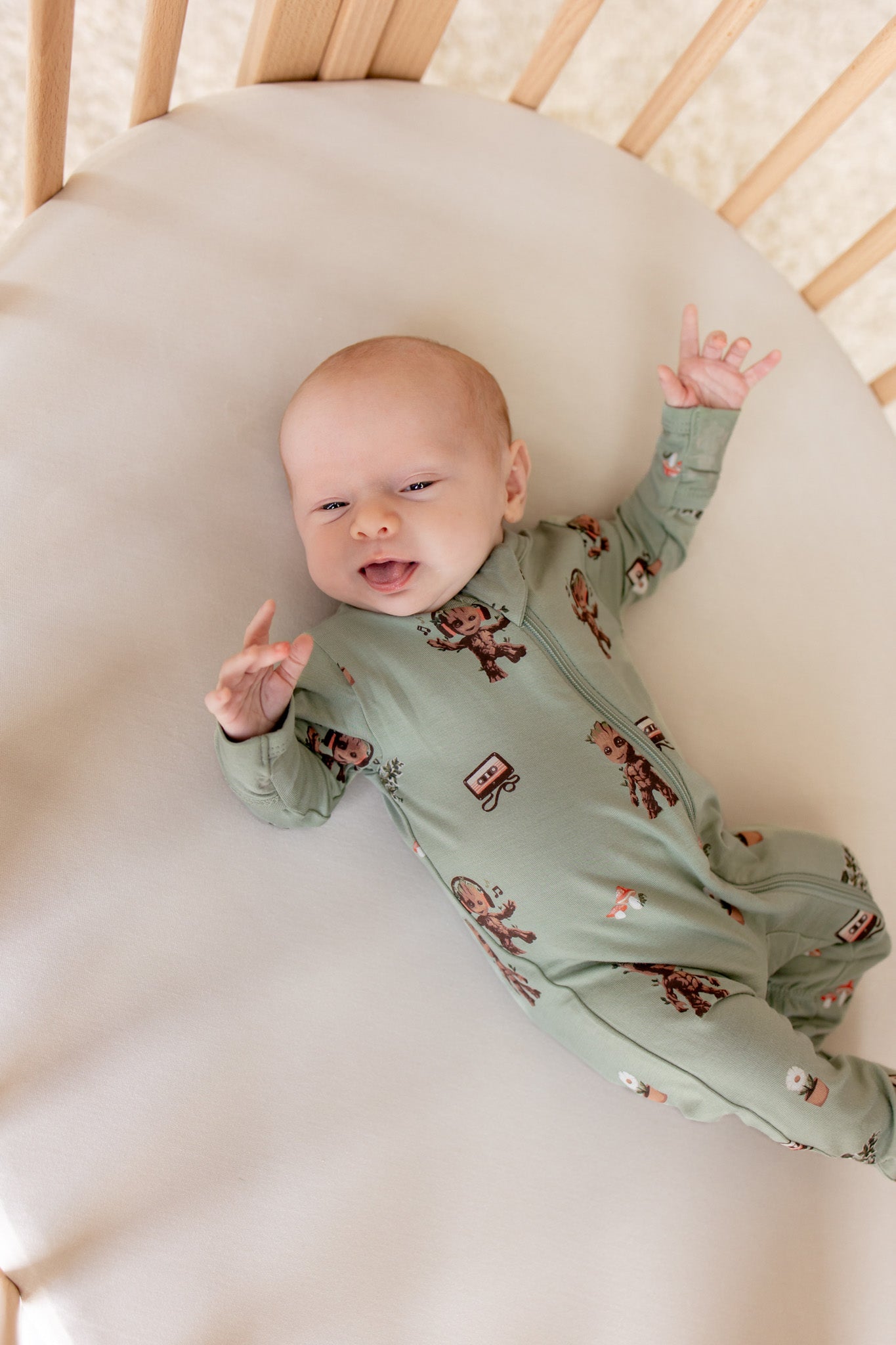 Baby lying in an oval cot wearing Green Zippered Footie in Groot print on a beige bassinet sheet