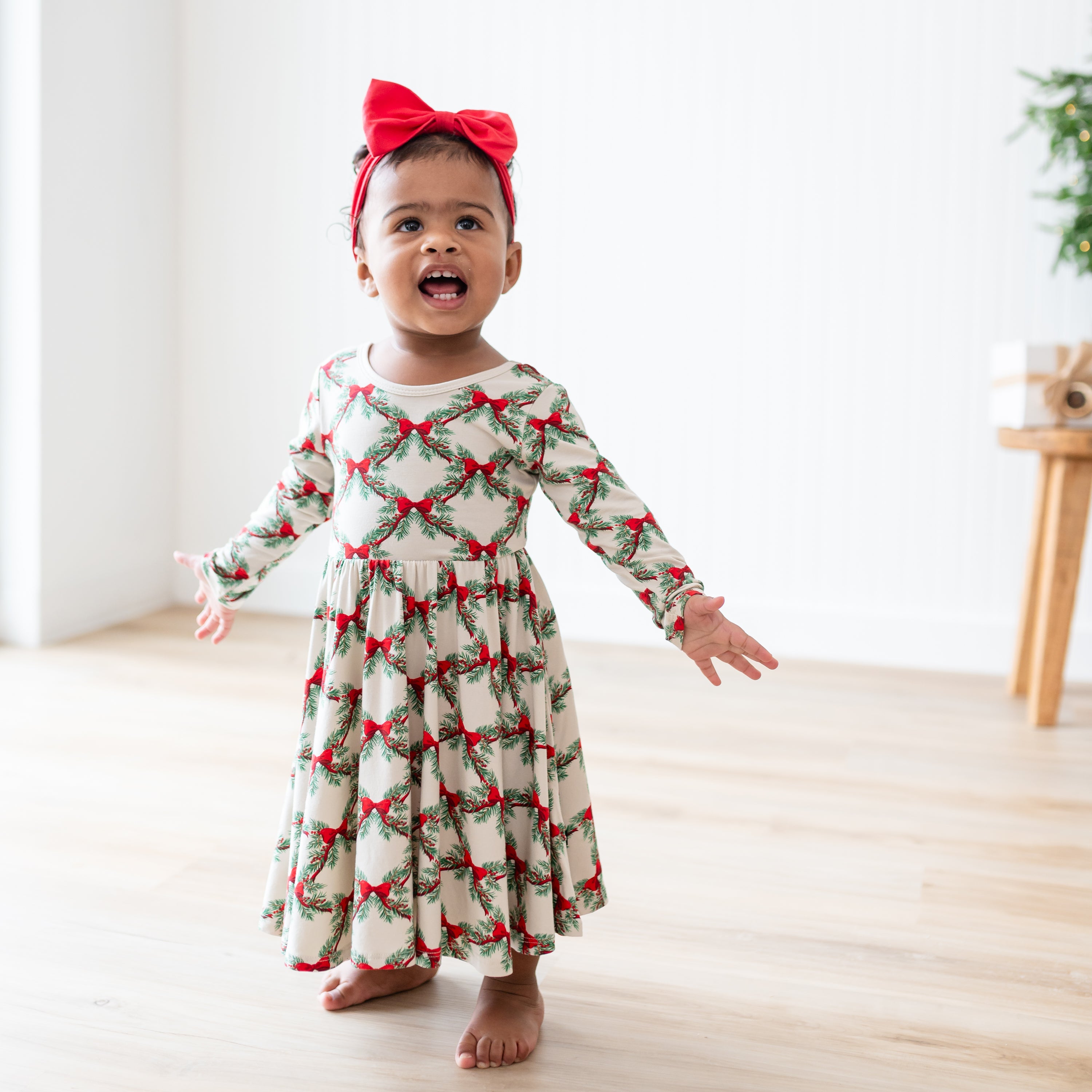Young toddler standing with her arms out wearing the Long Sleeve Twirl Dress in Holiday Bow paired with a Red bow headband