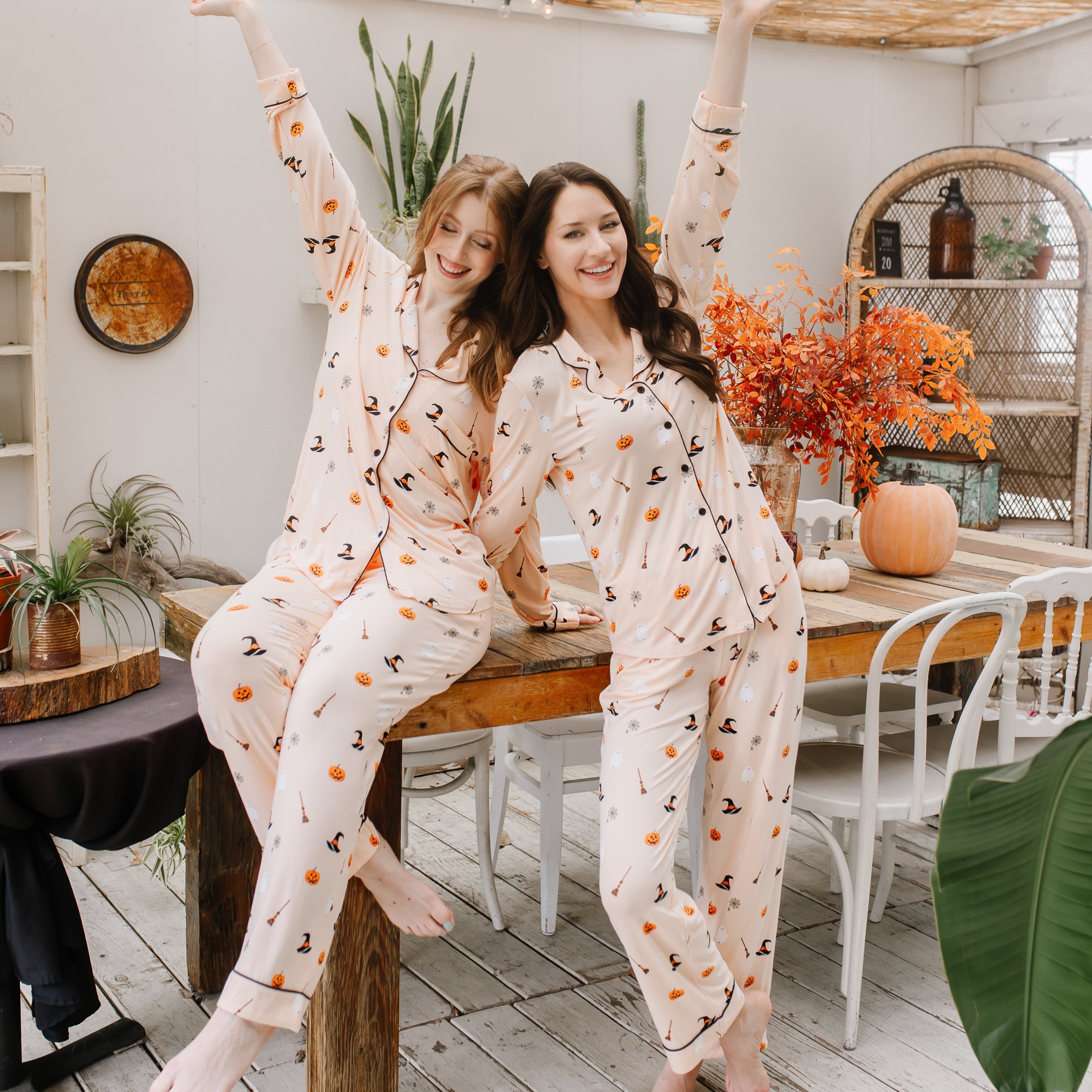 Two women in matching halloween printed pajamas sitting on a wooden table in a cozy room.
