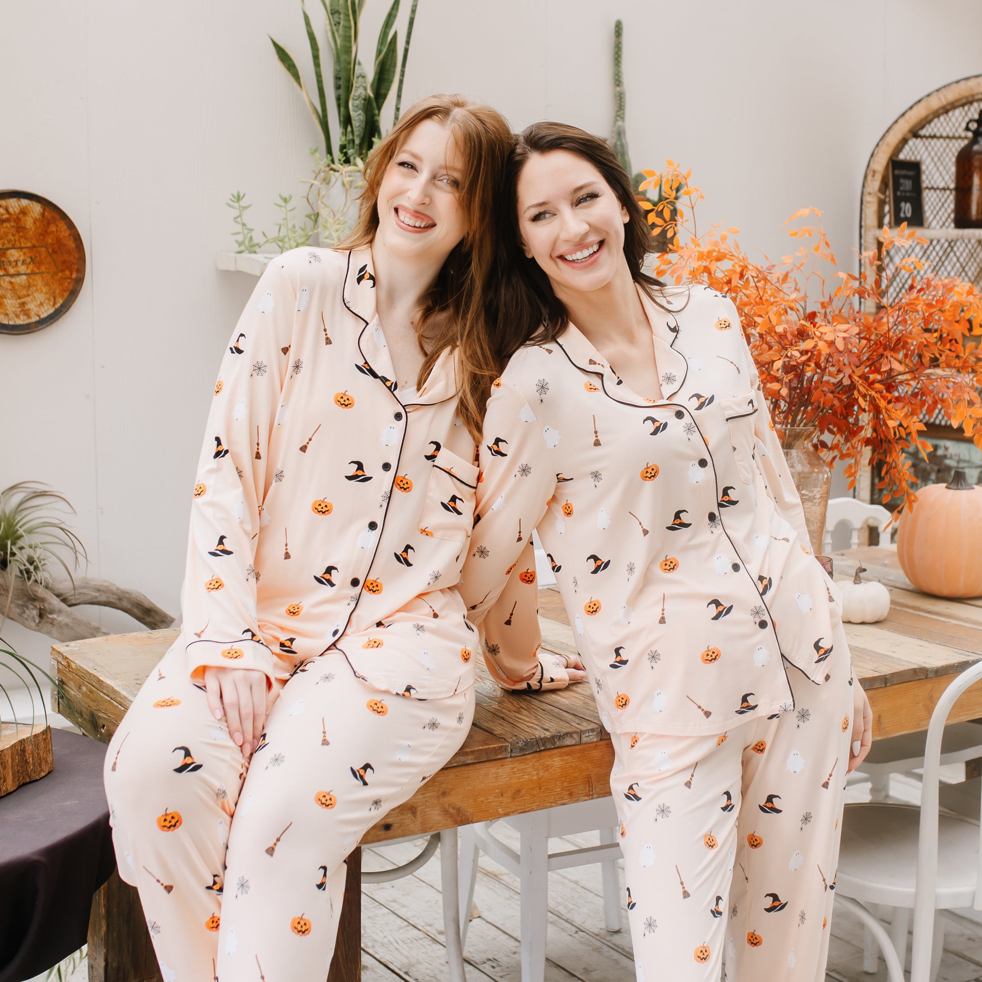 Two women wearing Halloween-themed pajamas sitting on a wooden stool indoors.
