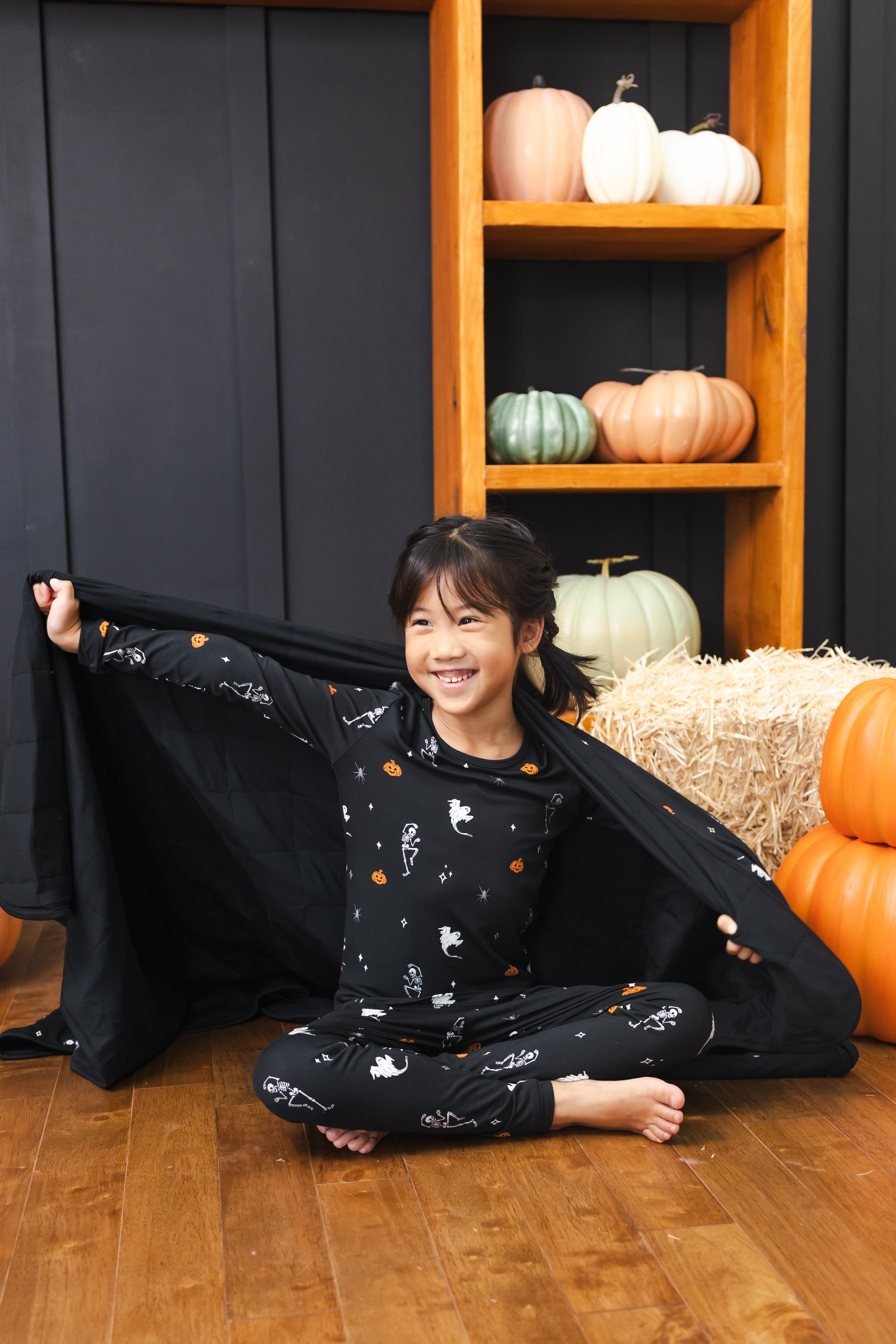 Girl holding Toddler Blanket in Halloween Friends 1.0 print wearing a similar print long sleeve pajama set sitting in front of Halloween decorations