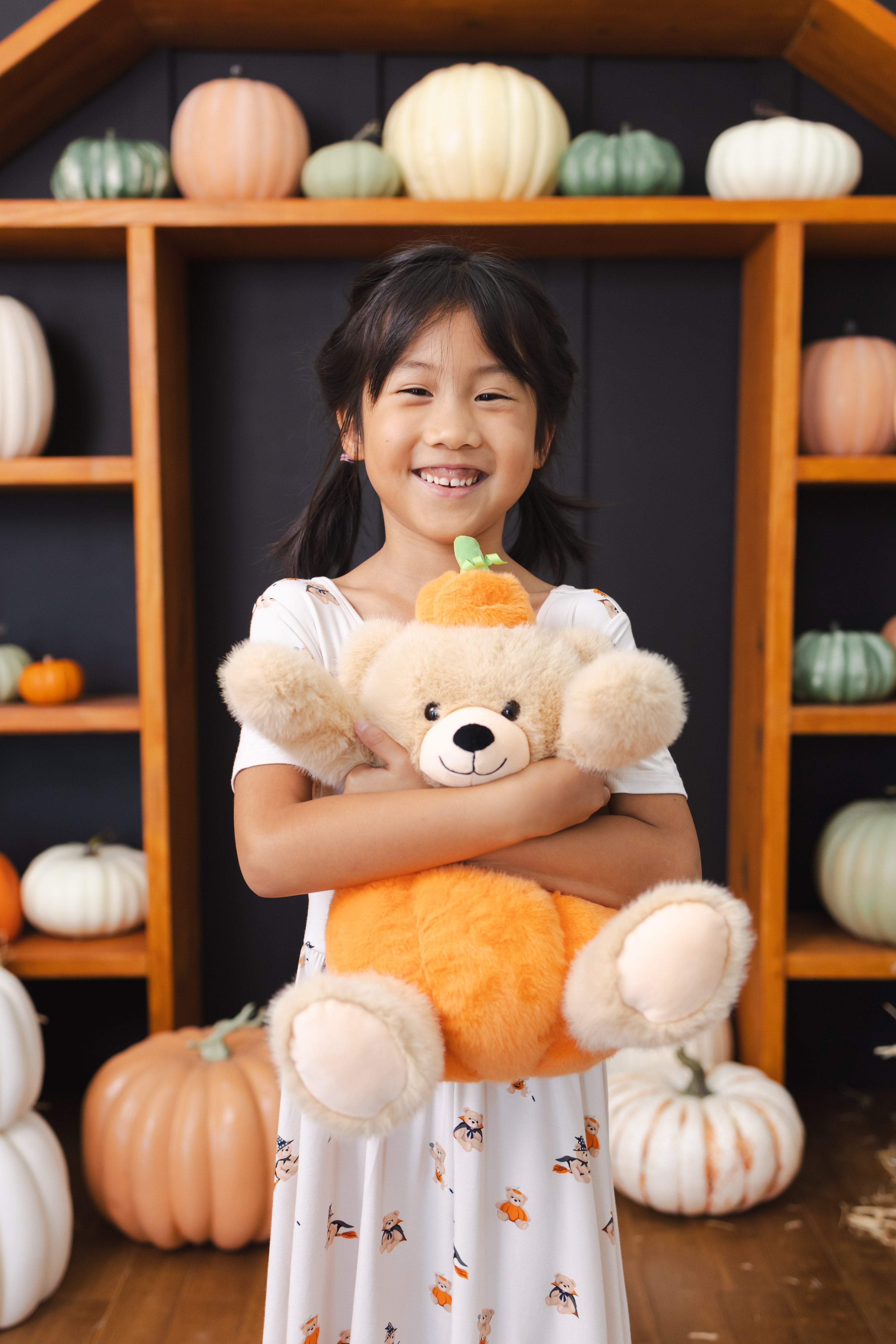 Girl standing with a stuffed teddy bear wearing Twirl Dress in Halloween Teddy print with pumpkin decorations around