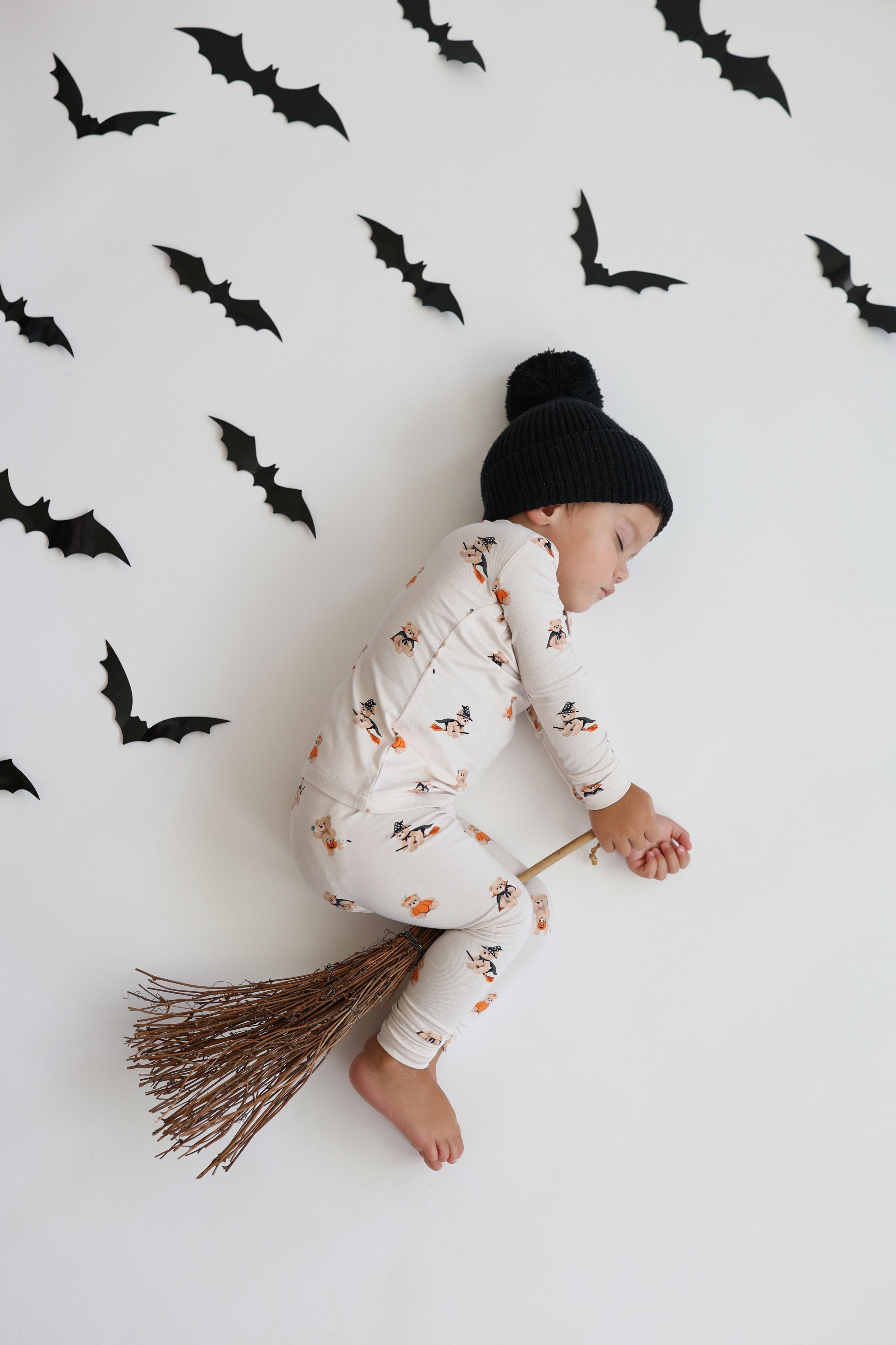 A baby wearing a black knit pom-pom beanie and a white long-sleeve romper with an orange and black witch/halloween print, posed on a broomstick against a white wall decorated with black bat cutouts.