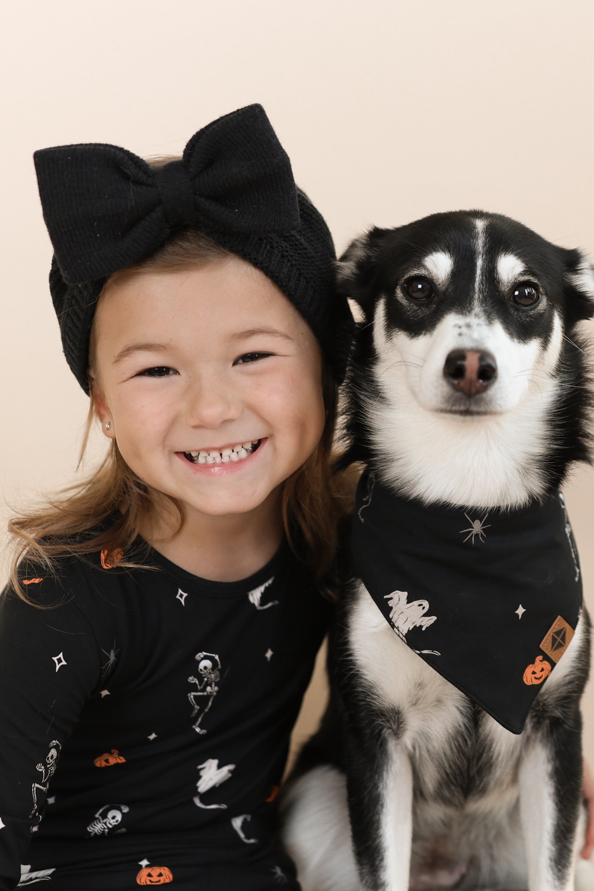 Black and White dog wearing Dog Bandana in Halloween Friends print sitting with a girl in similar print long sleeve pajama set on a beige background