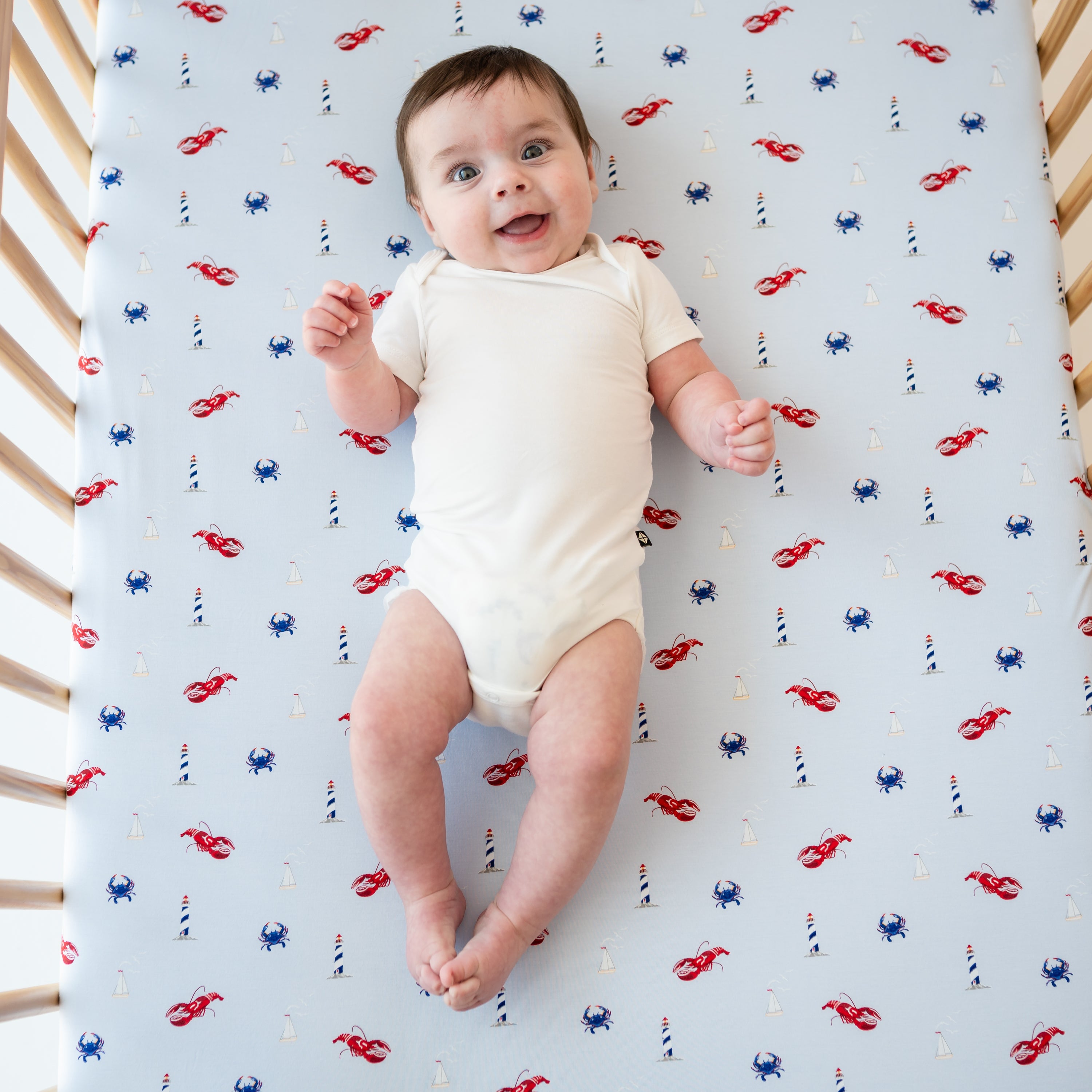 Smiling infant laying on top of a Crib Sheet in Harbor in a crib wearing the Cloud Short Sleeve Bodysuit