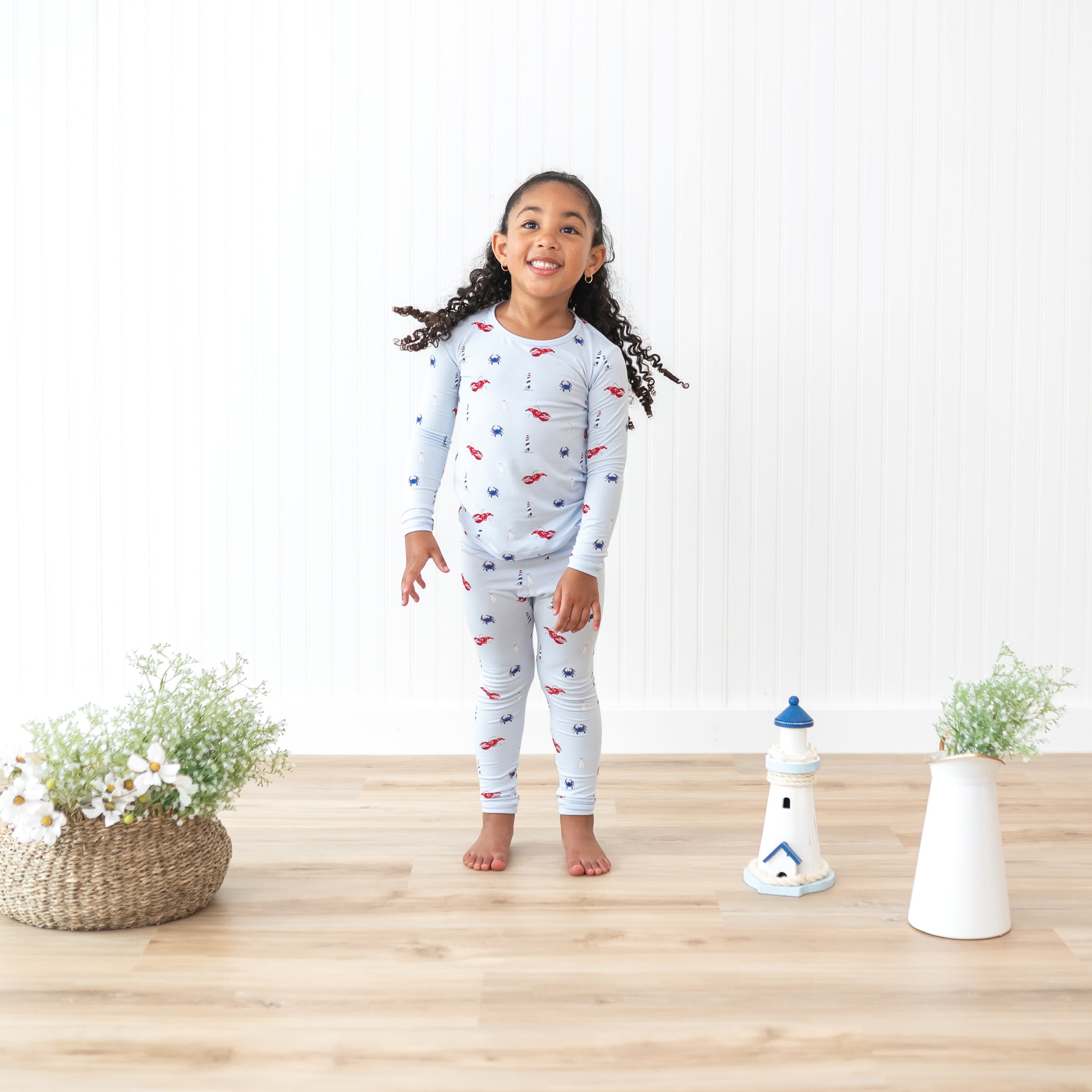Toddler model wearing the Long Sleeve Pajamas in Harbor beside potted flowers and a toy lighthouse
