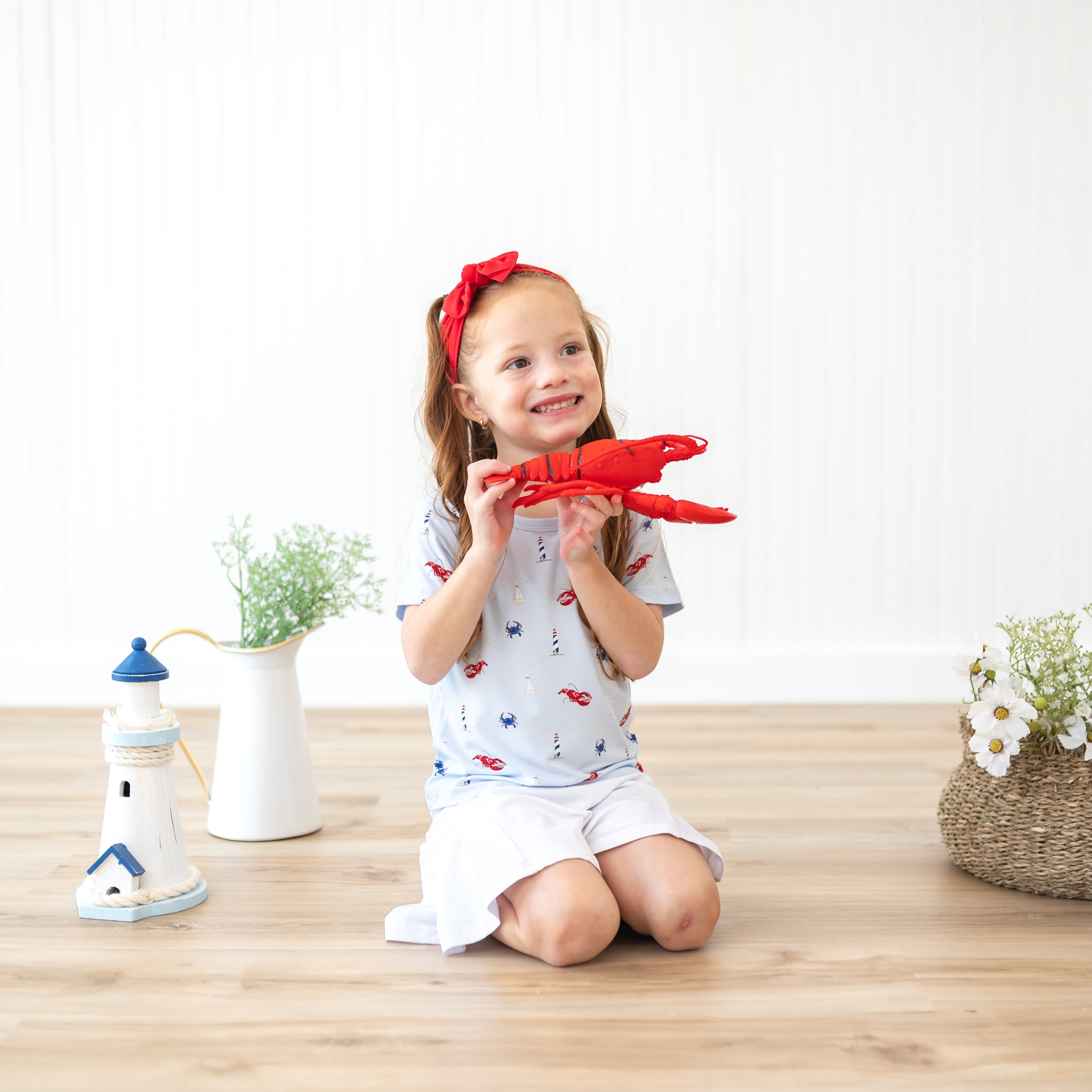 Toddler girl kneeling on the ground holding a stuffed toy lobster wearing the Toddler Basic Tee in Harbor, a white skirt and Knotted Bow Headband in Cardinal