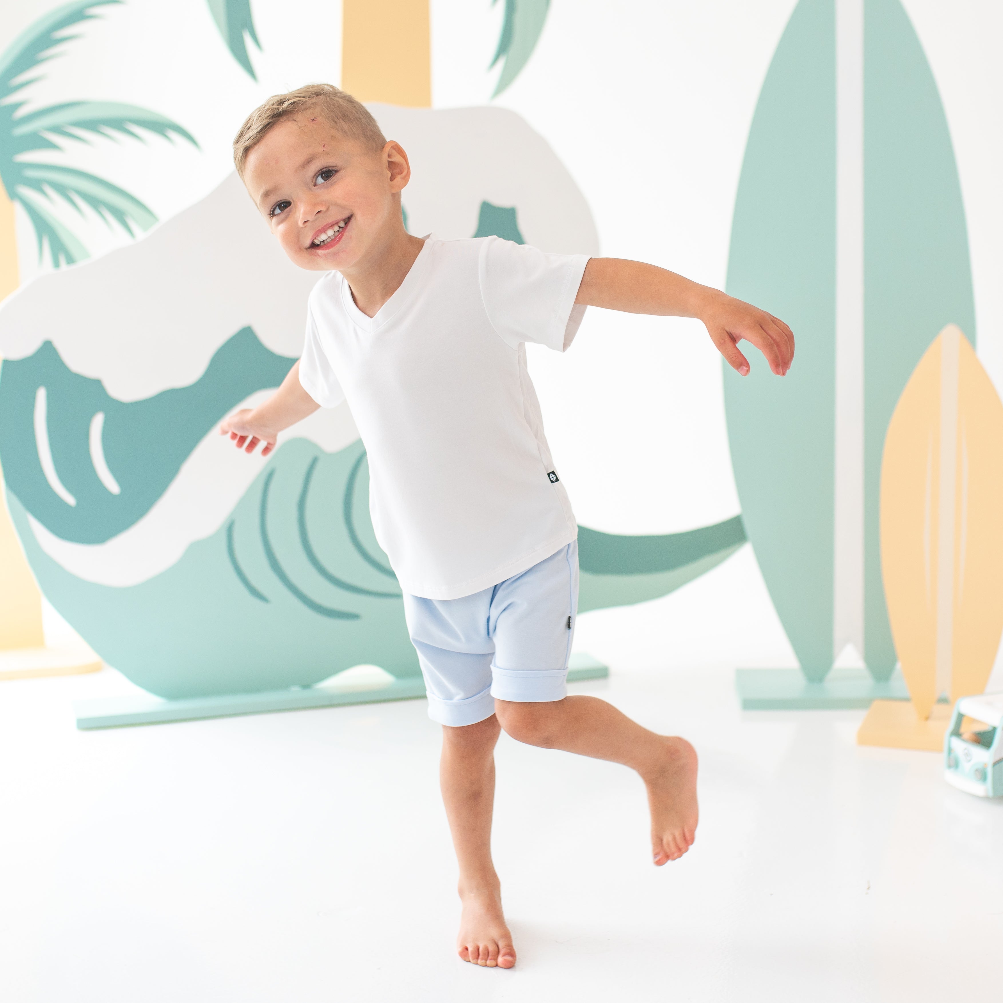 Young boy on one foot modeling the Harem Shorts in Mist paired with a Toddler V-Neck in Snow