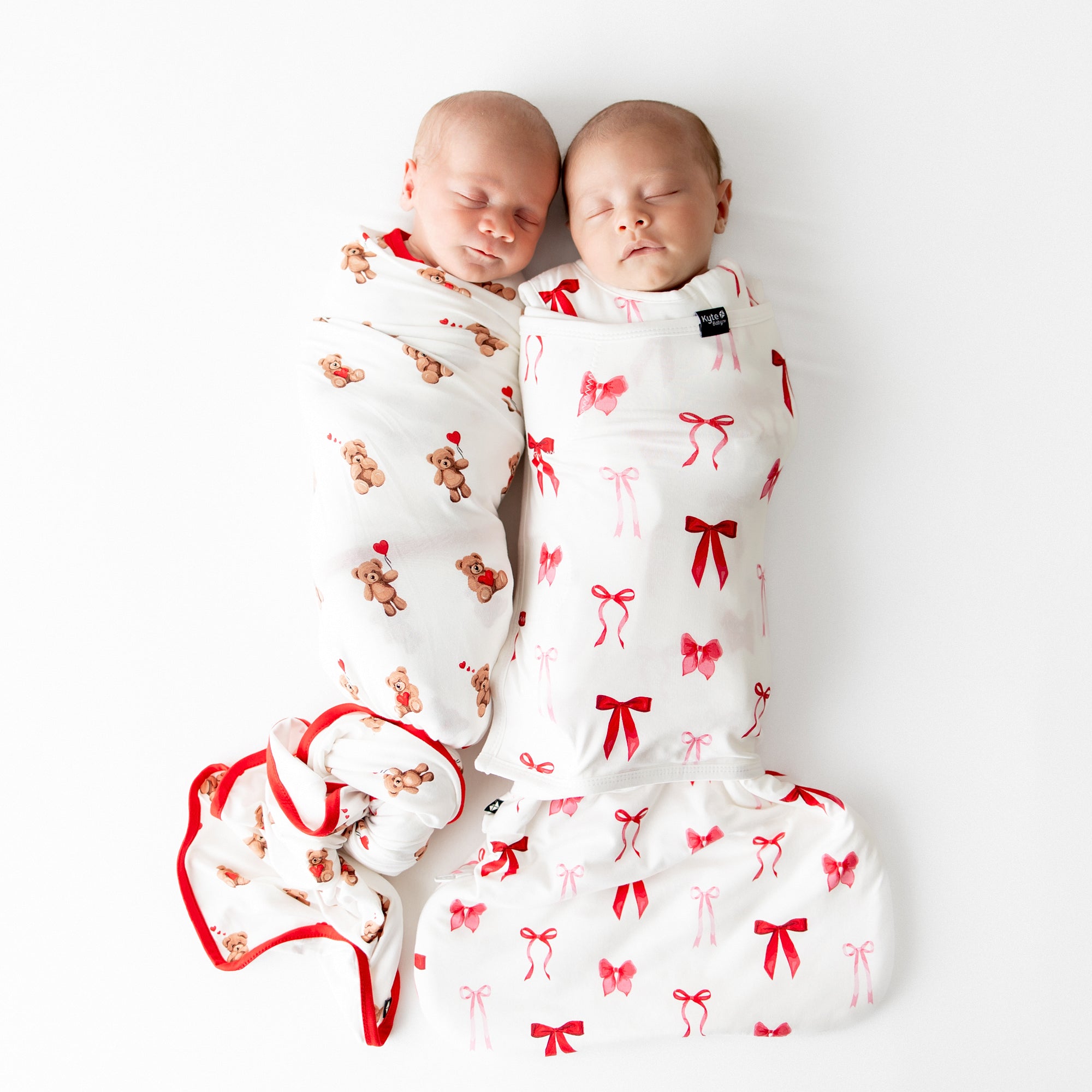 Two sleeping newborns laying side by side. One newborn wearing the Swaddle Blanket in Bear Hearts and the other wearing the sleep bag swaddler in small love bow