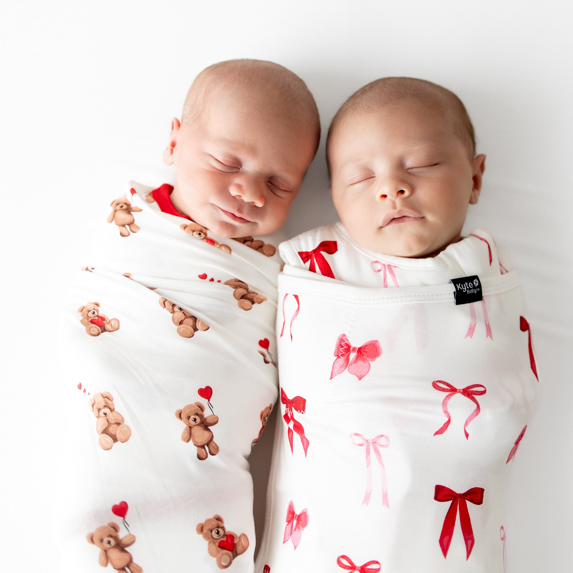 Two sleeping newborns laying side by side swaddled in the swaddle blanket in bear hearts and the Sleep Bag Swaddler in Small Love Bow 1.0