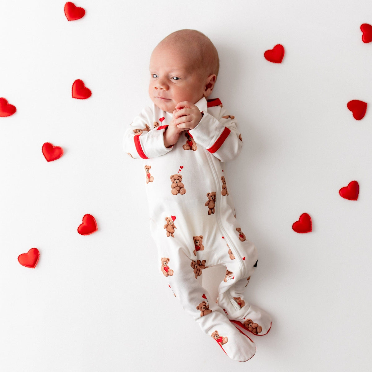 Newborn wearing the Zippered Footie in Bear Hearts laying on a white surface with small sewn red hearts around them