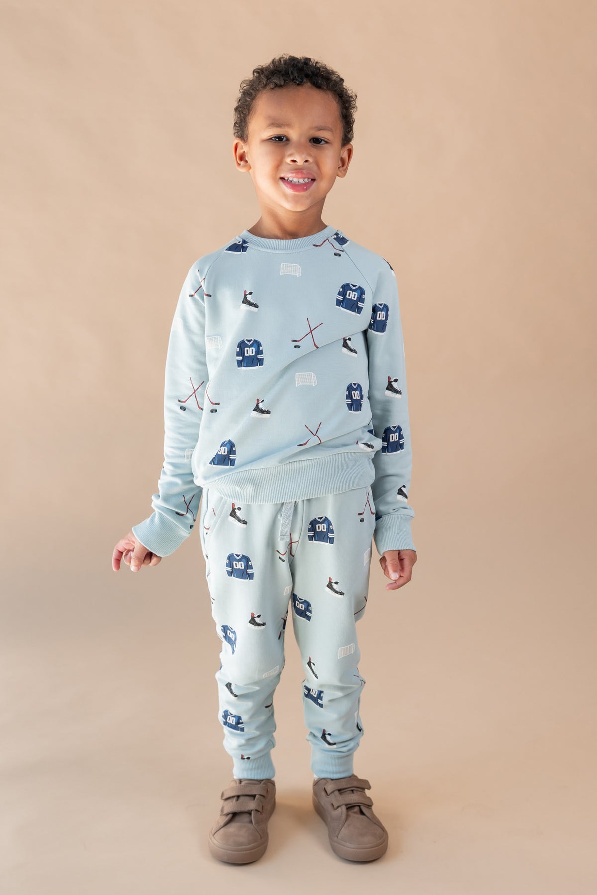 A young child wearing a light blue long-sleeve set with a dark blue and white ski pattern, paired with brown shoes, standing against a solid beige background.